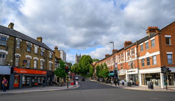 A street scene in a London neighbourhood with rows of brick terraced buildings, local shops, pedestrians, and a road junction under a partly cloudy sky.