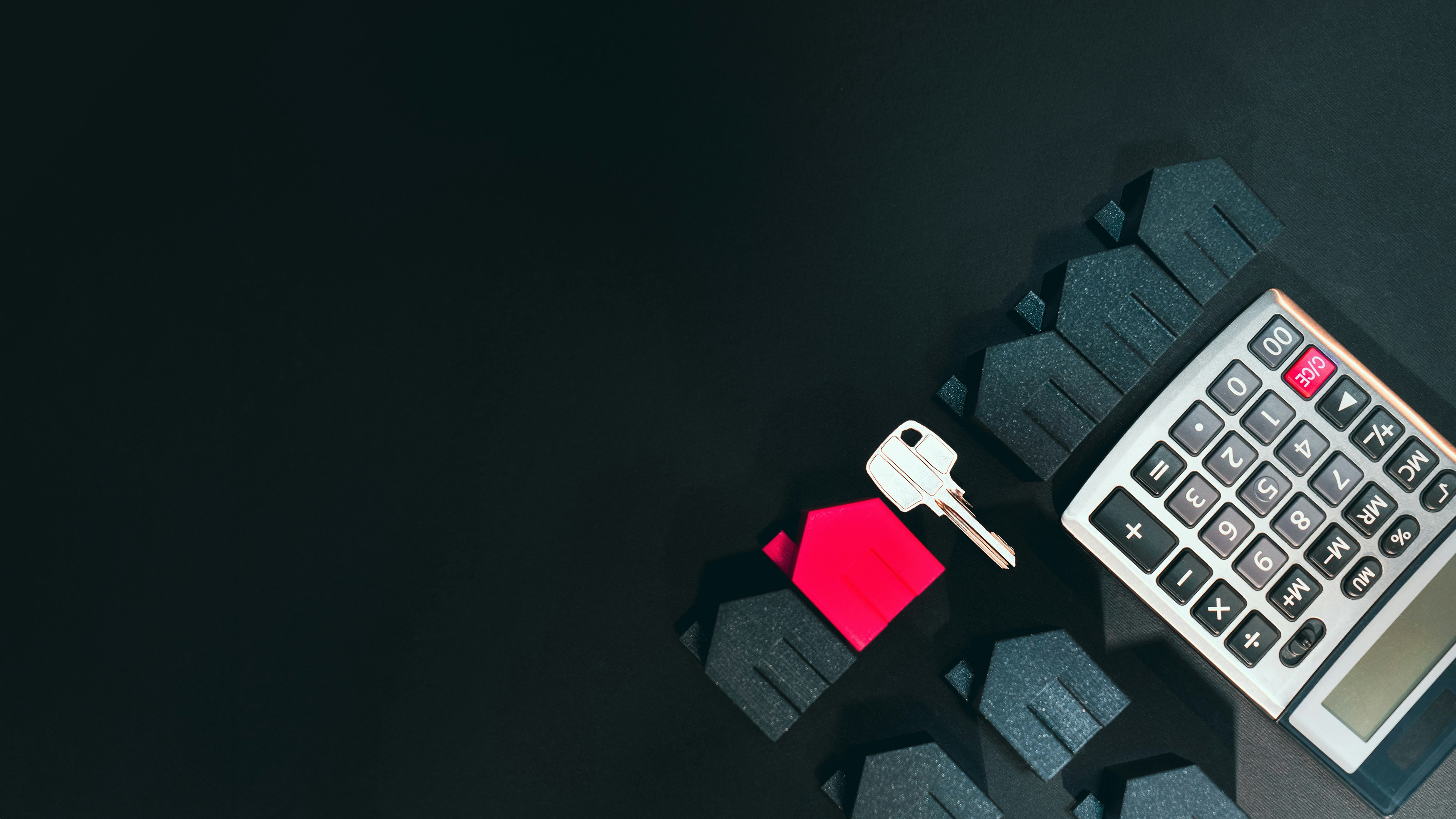 A collection of small house‑shaped models is arranged on a dark surface. One house is colored red, standing out among the black houses. A silver key rests on top of the red house, symbolizing ownership or access. To the right, a calculator suggests a financial or real estate context such as mortgage calculation, property investment, or home buying.