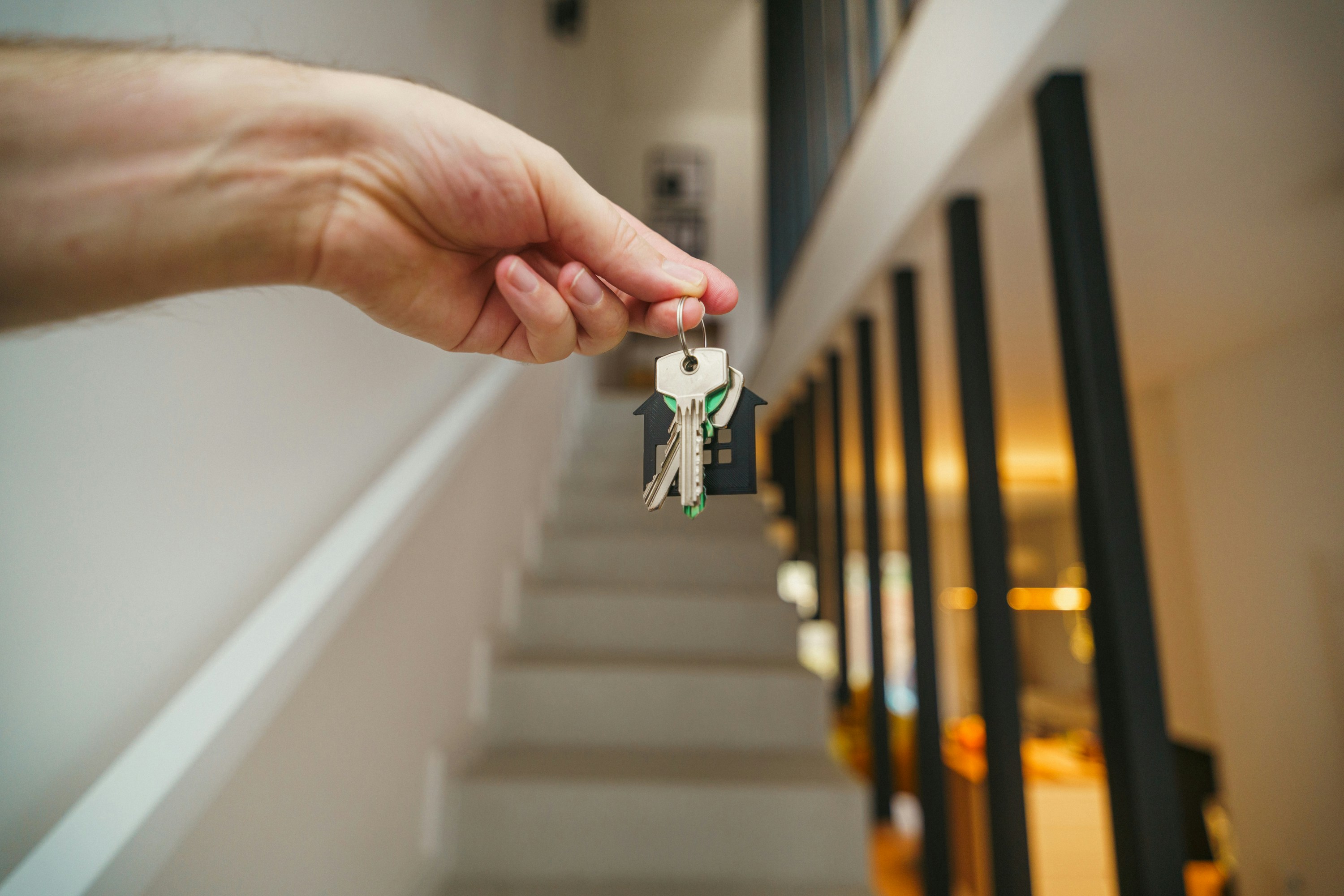 A close‑up shows a hand holding a set of house keys attached to a black house‑shaped key fob. The background reveals a modern interior with vertical black railings along a staircase and warm lighting, suggesting a newly acquired or rented property.