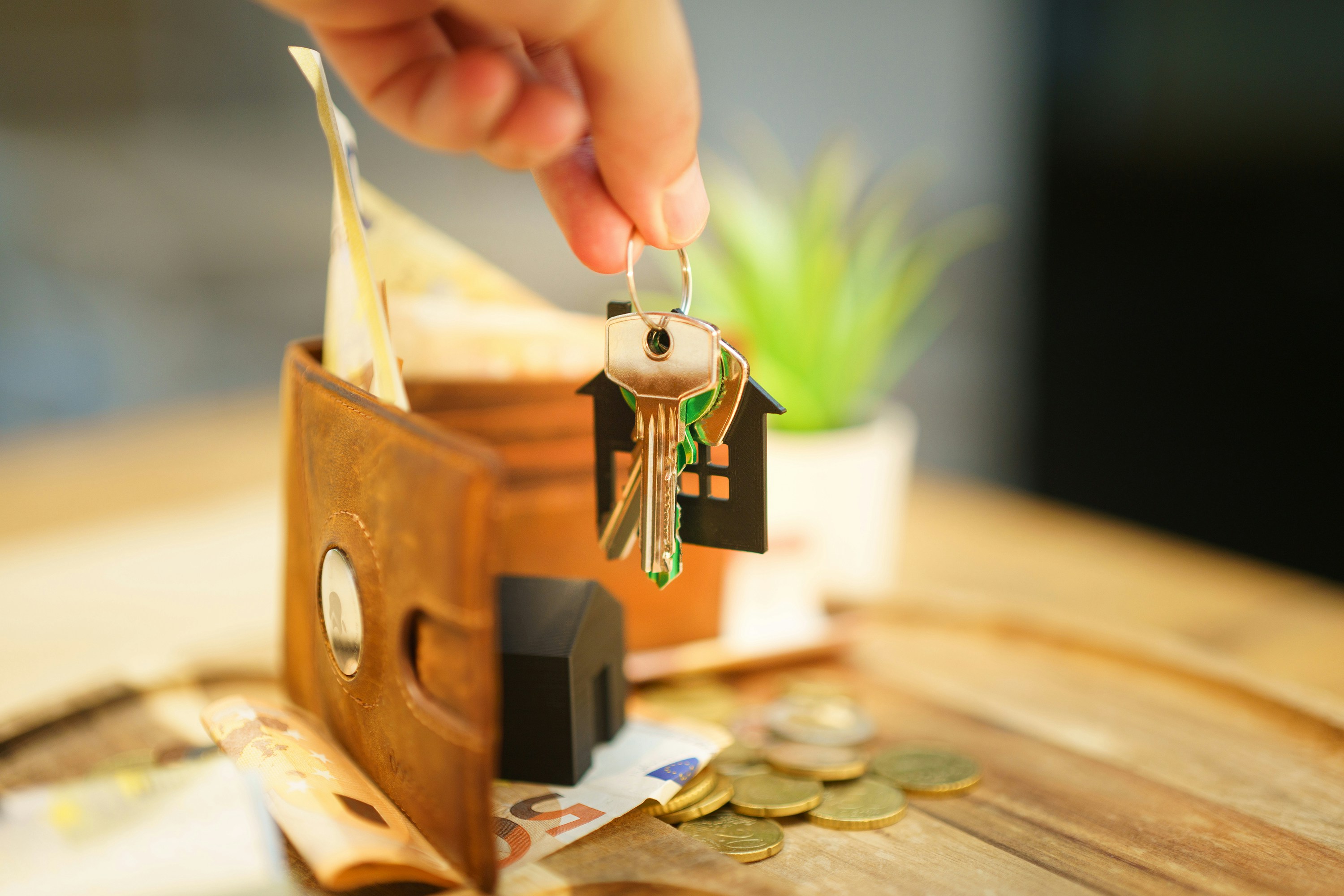 A hand holds a keychain with house-shaped fobs and keys above a wooden surface displaying a brown leather wallet with euro banknotes, a small black house model, and scattered euro coins. A blurred green plant in a white pot sits in the background, evoking themes of homeownership, real estate investment, and financial planning.