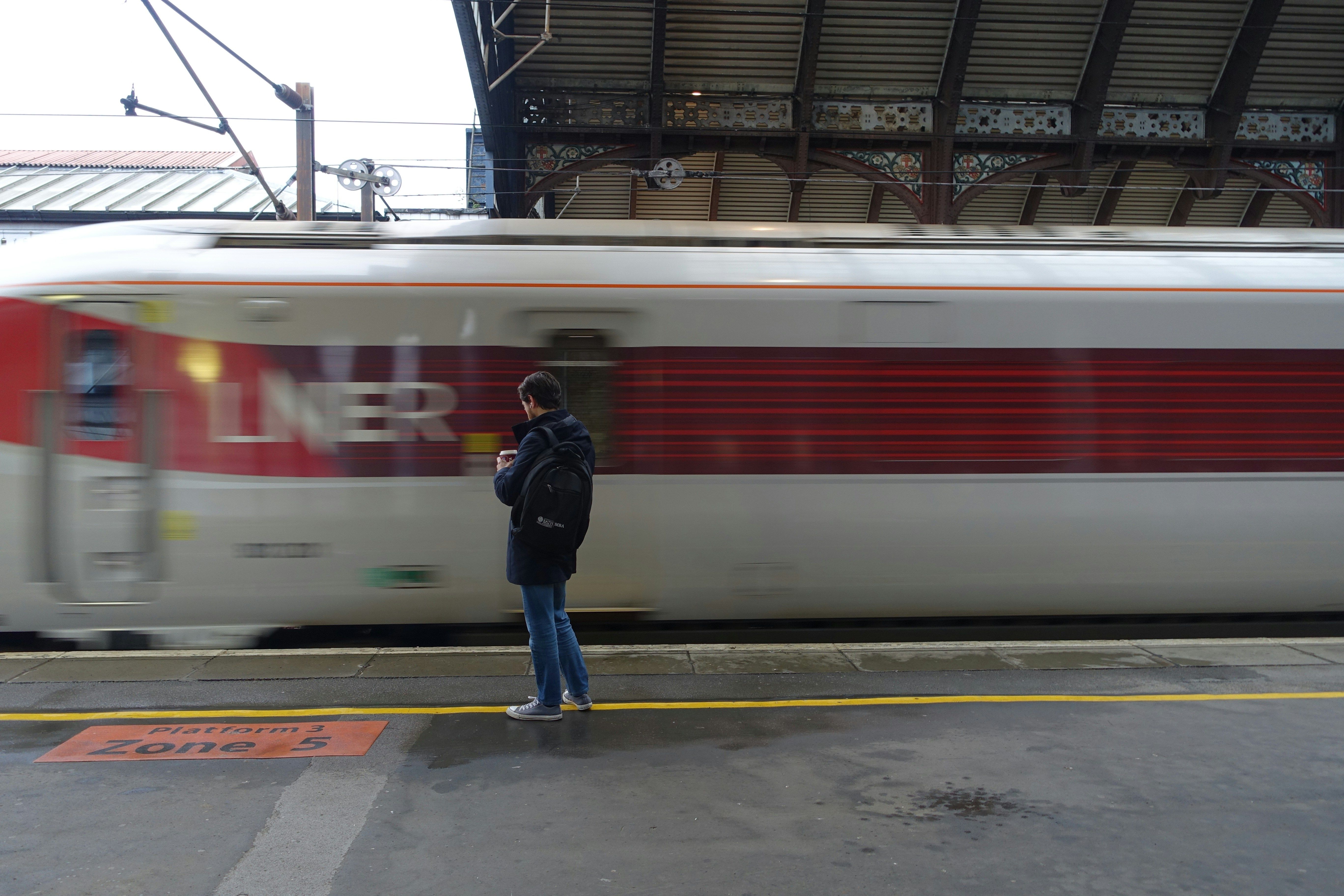 "Person on a train platform as a fast-moving LNER train passes by. Sign reads 'Platform 3 Zone 5.