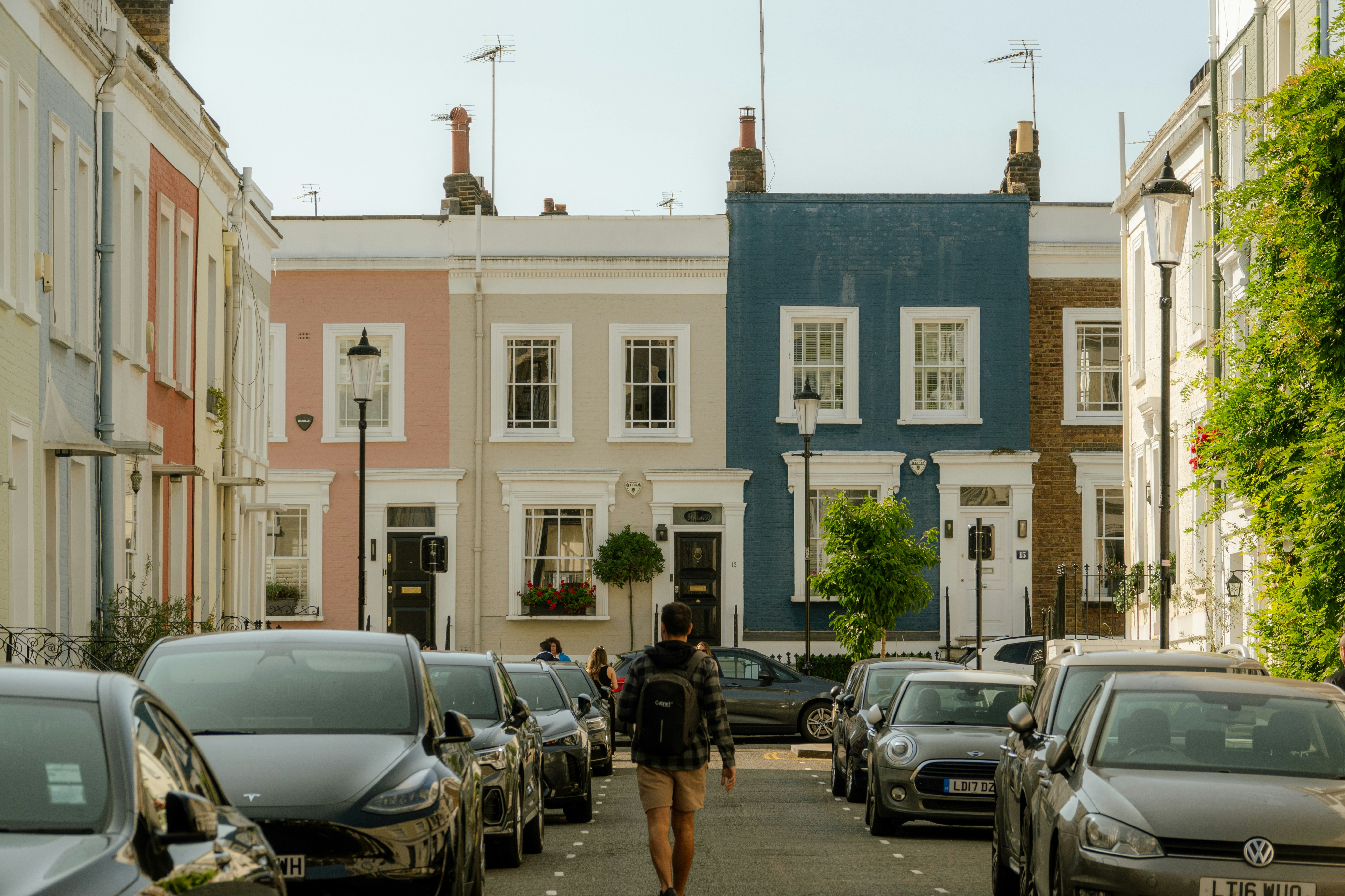A picturesque residential street features pastel-painted terraced houses with sash windows, black front doors, and flower boxes. Parked cars, including a Tesla and a Mini Cooper, line both sides of the street. A person wearing a backpack and shorts walks down the center. Traditional street lamps and rooftop antennas add to the charm of this quiet urban neighborhood.