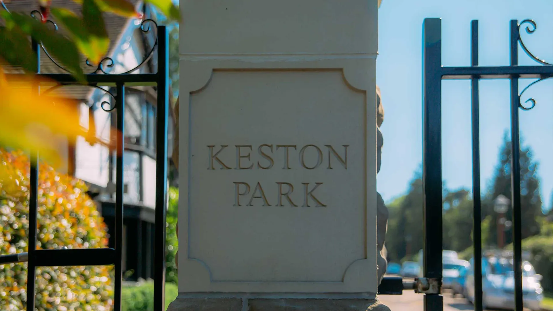 A stone pillar engraved with the words 'Keston Park' stands beside ornate black wrought iron gates with decorative scrollwork. Behind the entrance, residential houses, greenery, and parked cars line the street under a bright blue sky.