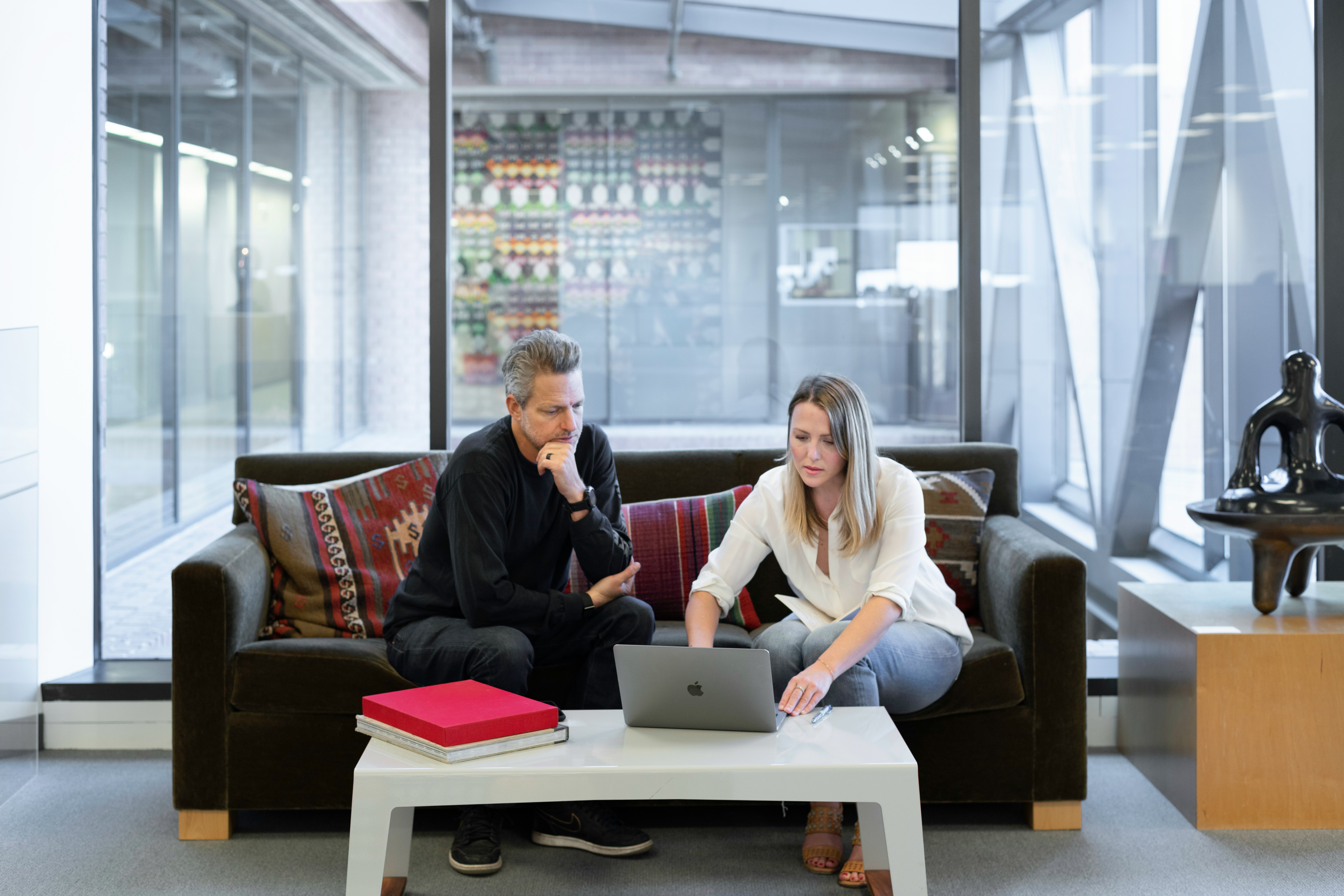 Two individuals sit on a dark brown couch in a modern office lounge, focused on a laptop placed on a white coffee table. One wears a black outfit and observes, while the other, dressed in a white blouse and jeans, engages with the screen. The table also holds two large books, one red. Behind them, a glass wall reveals colorful wall art and contemporary architecture, suggesting a clean, professional, and collaborative environment.
