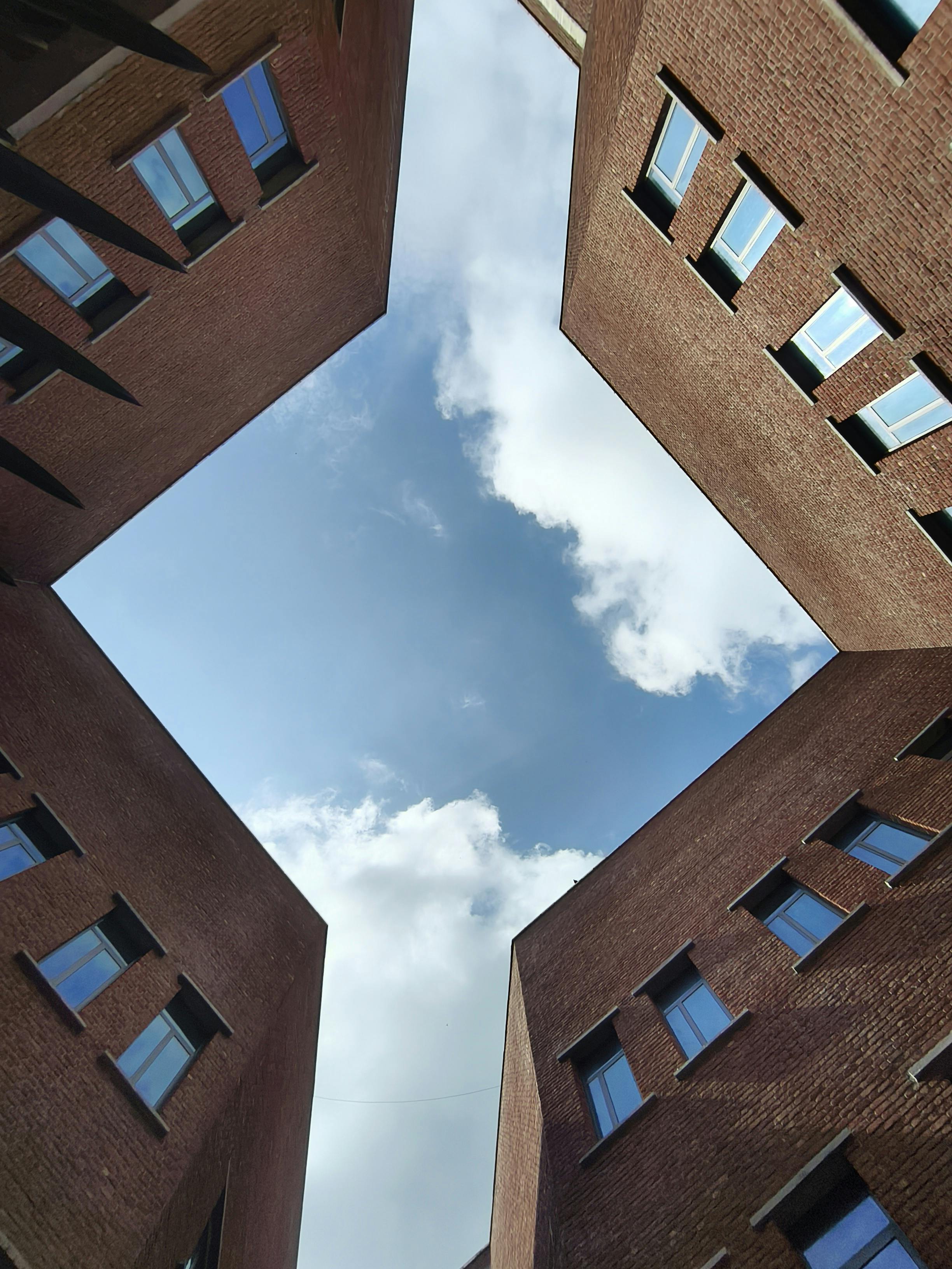 Looking up from the center of a courtyard, four brick buildings form a symmetrical frame around a partly cloudy sky. Each building features consistent brickwork and multiple windows, creating a geometric composition that contrasts the structured architecture with the openness of the sky.