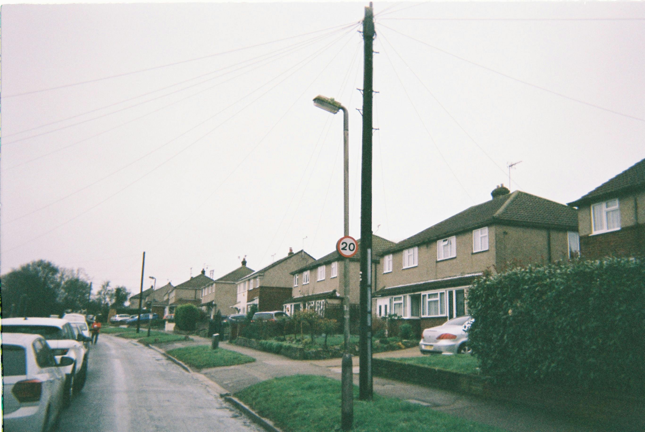 A suburban street lined with semi-detached brick houses features tiled roofs, small front gardens, and driveways. Several cars are parked along the wet road and in driveways, suggesting recent rain. A streetlamp and utility pole with overhead wires stand prominently in the foreground, with a circular sign indicating a 20 mph speed limit. The overcast sky adds to the quiet residential atmosphere.