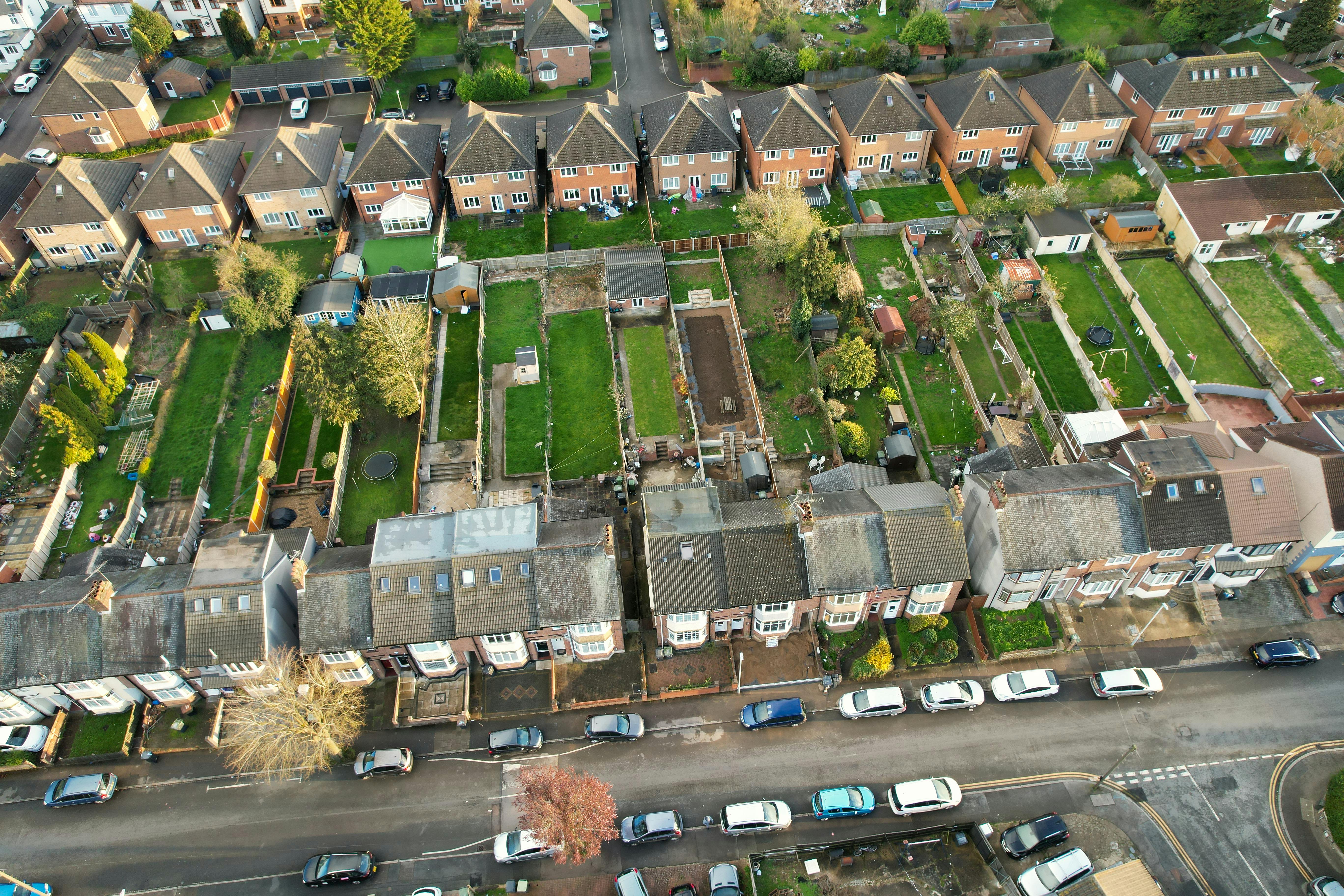 An aerial view of a residential area showing two rows of houses with backyards. The lower row features smaller yards, while the upper row includes larger lawns, garden sheds, and landscaping. Streets border the neighborhood, with cars parked along the curb, illustrating a well-organized suburban layout.