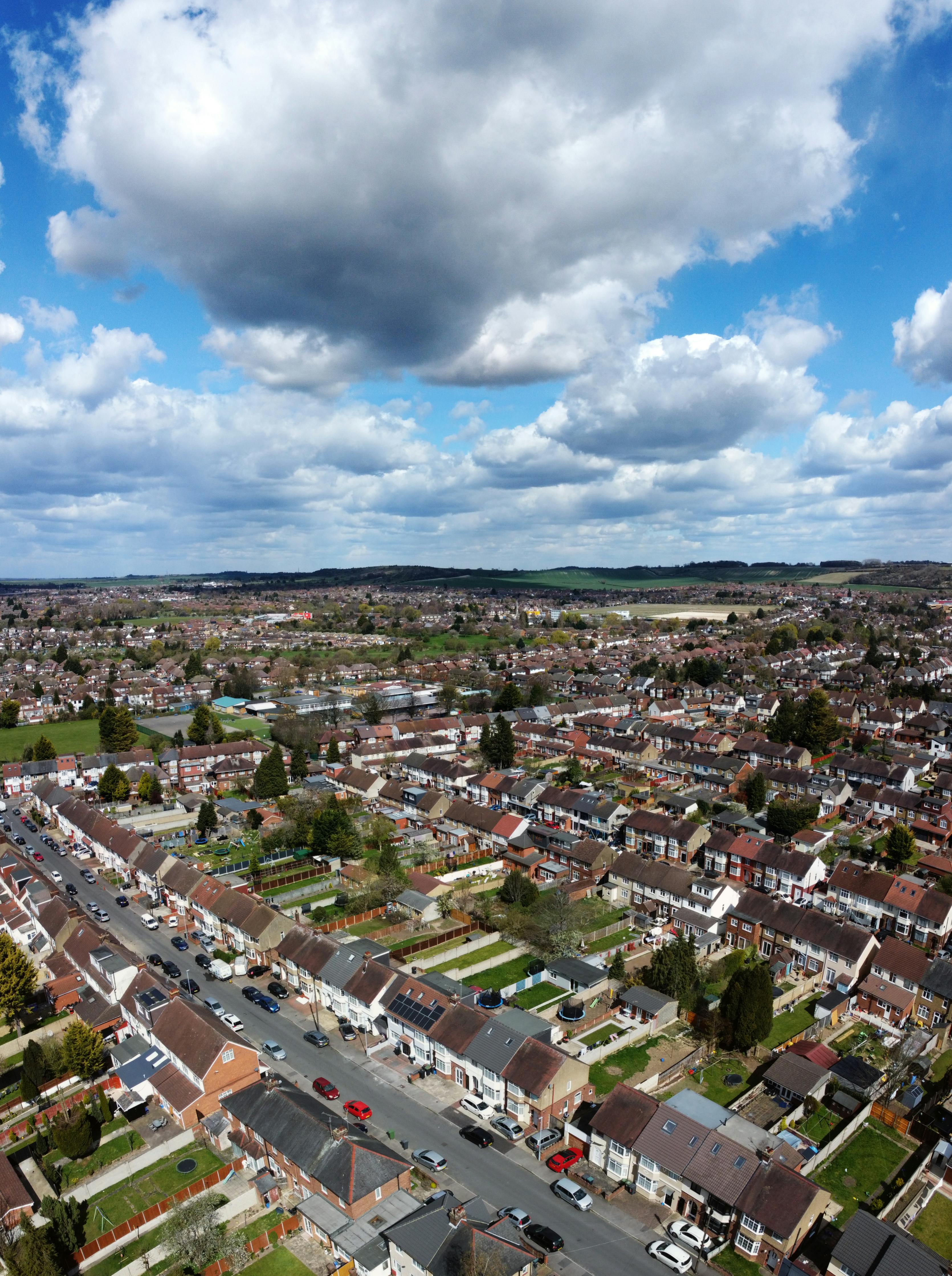 An aerial view of a well-organized suburban area shows rows of closely packed houses with small gardens and brown or red roofs, some fitted with solar panels. Streets lined with parked cars weave through the neighborhood, which transitions into green fields and rolling hills in the distance. Large, fluffy clouds fill the sky, adding drama to the peaceful urban-rural landscape.