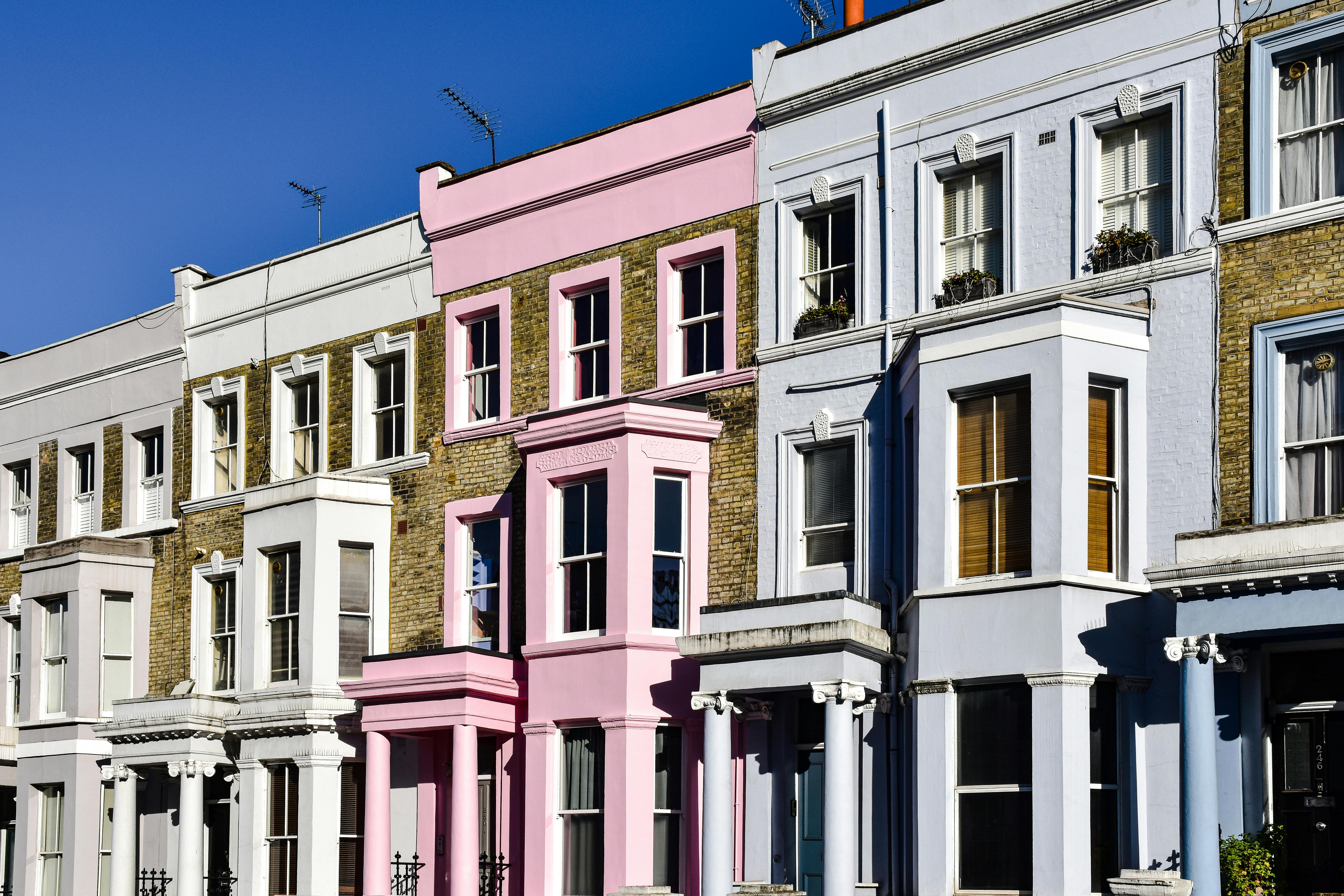 A row of pastel-colored terraced houses in shades of white, pink, blue, and lavender lines an urban street. Each house features bay windows, decorative cornices, and columned entrances, reflecting Victorian architectural influence. The buildings are closely packed, creating a vibrant and picturesque residential scene.