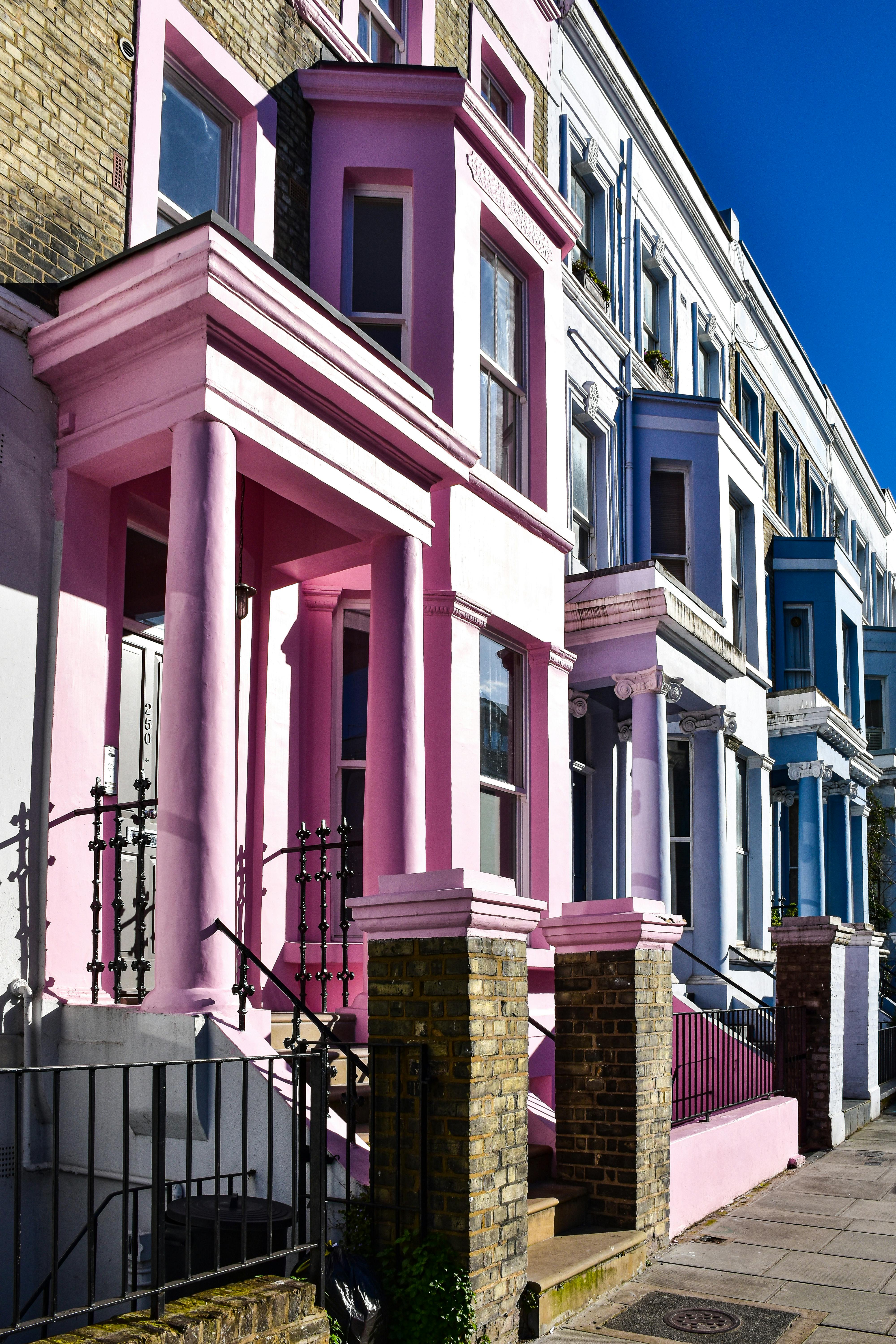 A row of vibrant terraced houses painted in pink, lavender, white, and blue lines an urban street. Each house features classical architectural details like columns, cornices, and bay windows. The house in the foreground, painted bright pink, displays the number “250” on its door. Brick pillars with iron railings and a clean sidewalk enhance the cheerful and distinctive streetscape.