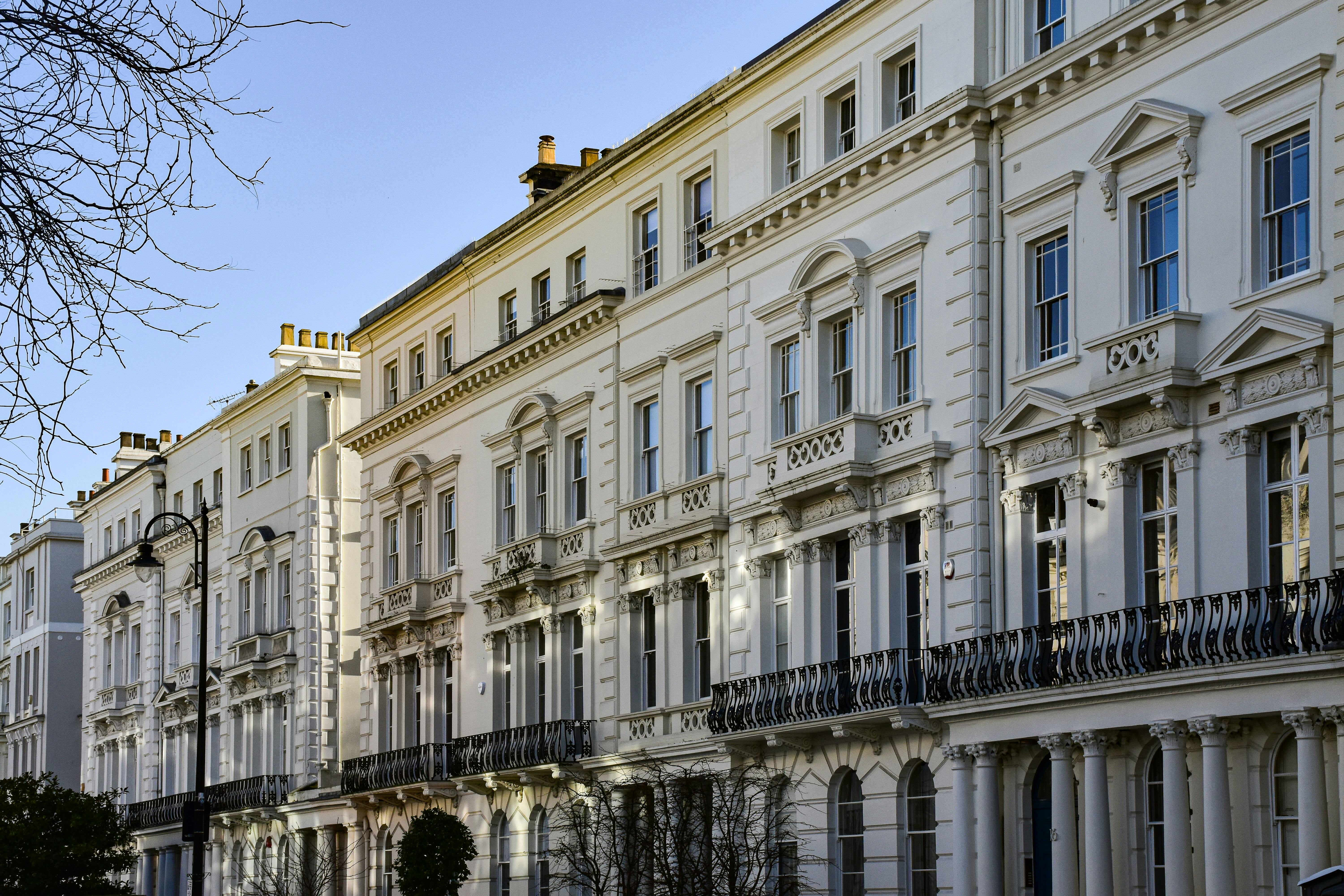 A row of multi‑story terraced houses painted in light cream features classical detailing, including decorative cornices, pilasters, arched windows, and wrought iron balconies. The facades are symmetrically arranged with intricate moldings. In the foreground, a black streetlamp and bare tree branches suggest a winter or early spring setting, complementing the refined, historic urban architecture.