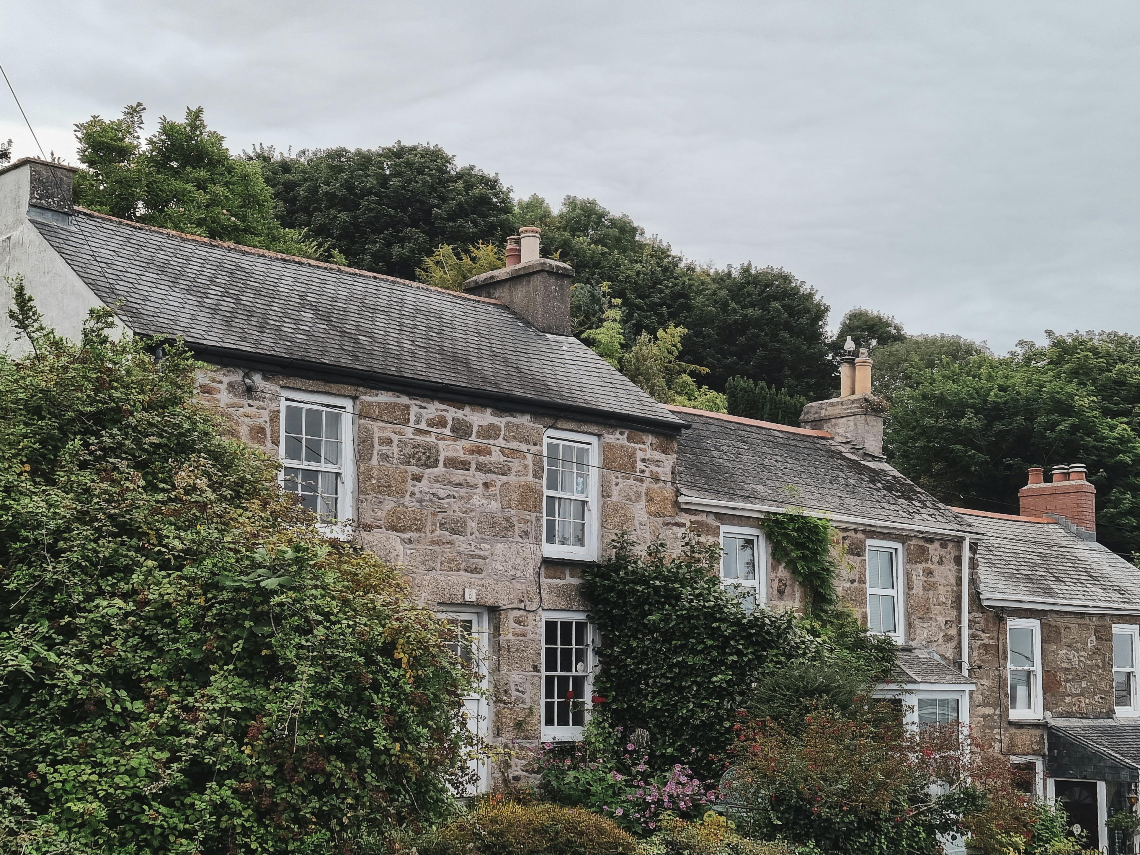 A row of traditional stone houses with slate roofs and white-framed windows sits amid lush greenery and blooming plants. Each house features a chimney, and dense trees in the background rise beneath an overcast sky, creating a tranquil countryside atmosphere.