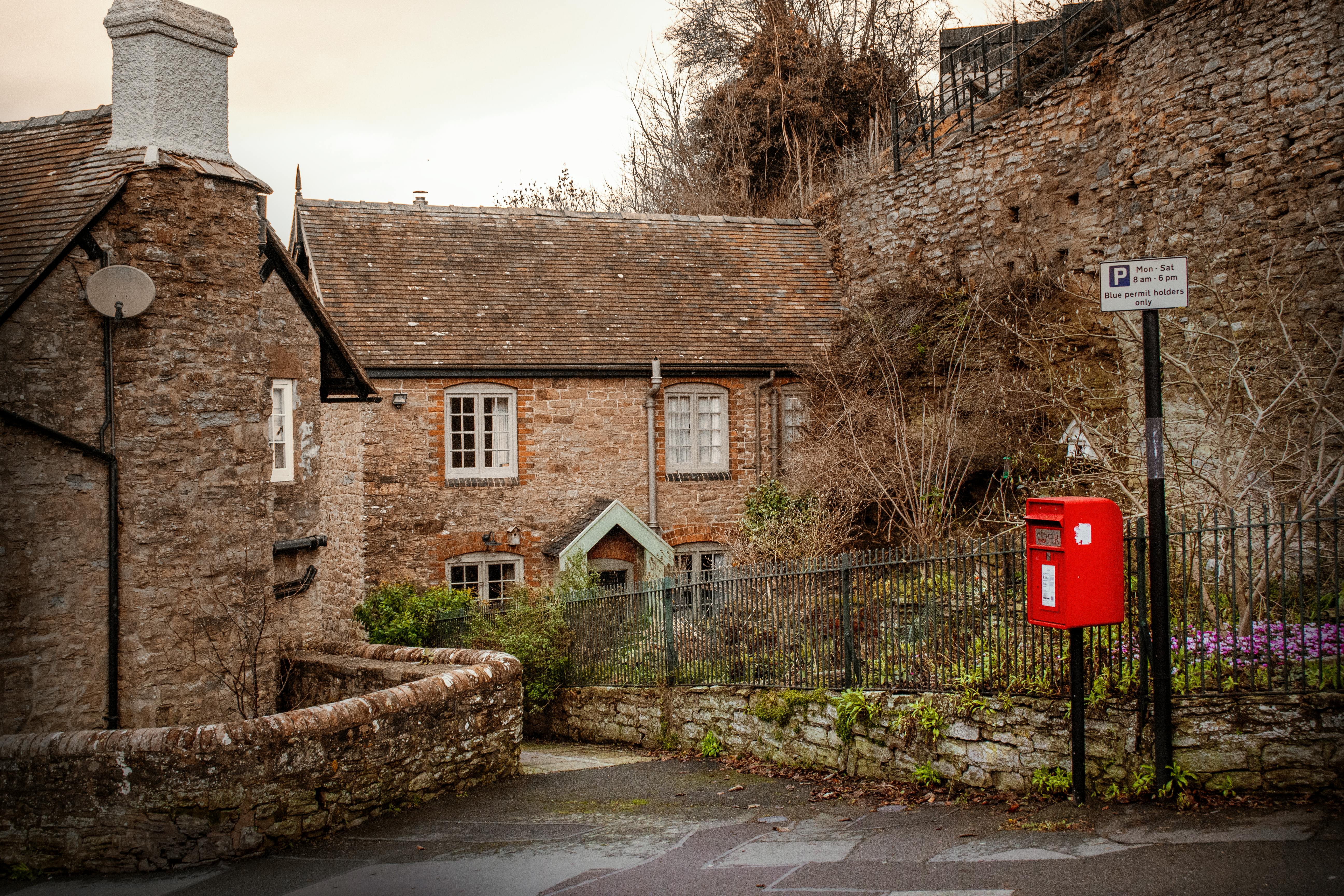 A quaint village scene shows traditional stone buildings with tiled roofs, white‑framed windows, and iron fencing. A red post box mounted on a black pole stands near the sidewalk, with a parking sign above it that reads “Mon – Sat, 8am – 6pm, Blue permit holders only.” A small garden with greenery and purple flowers adds color, while a satellite dish is mounted on one of the buildings. The overall setting highlights the charm of an English village street.