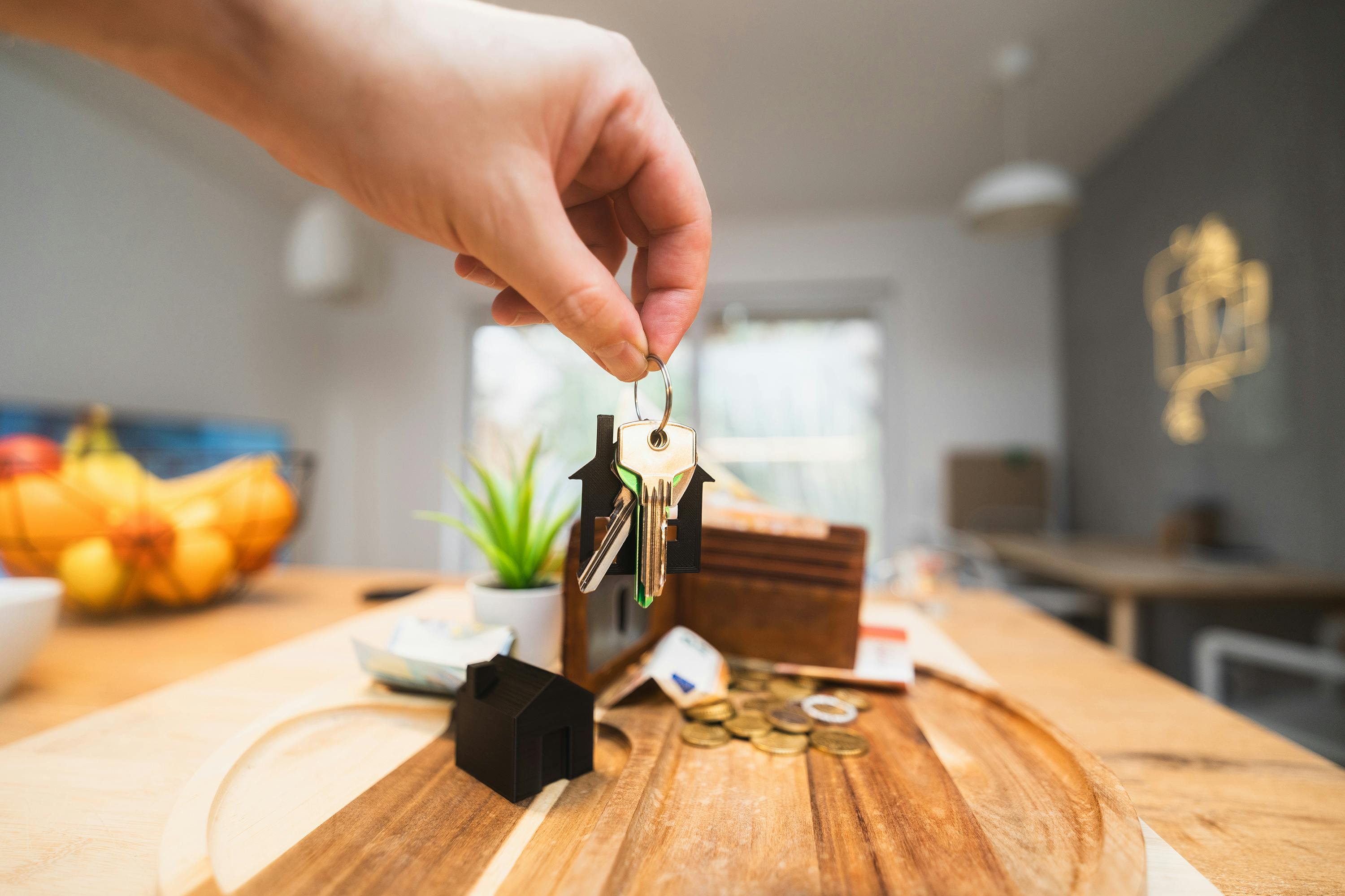 A close-up of a hand holding house keys with a house-shaped keychain. In the background, a wooden table displays a small black house model, coins, paper currency, a wallet, a potted plant, and a fruit basket. Natural light filters into the modern kitchen or dining space, evoking themes of homeownership and financial planning.