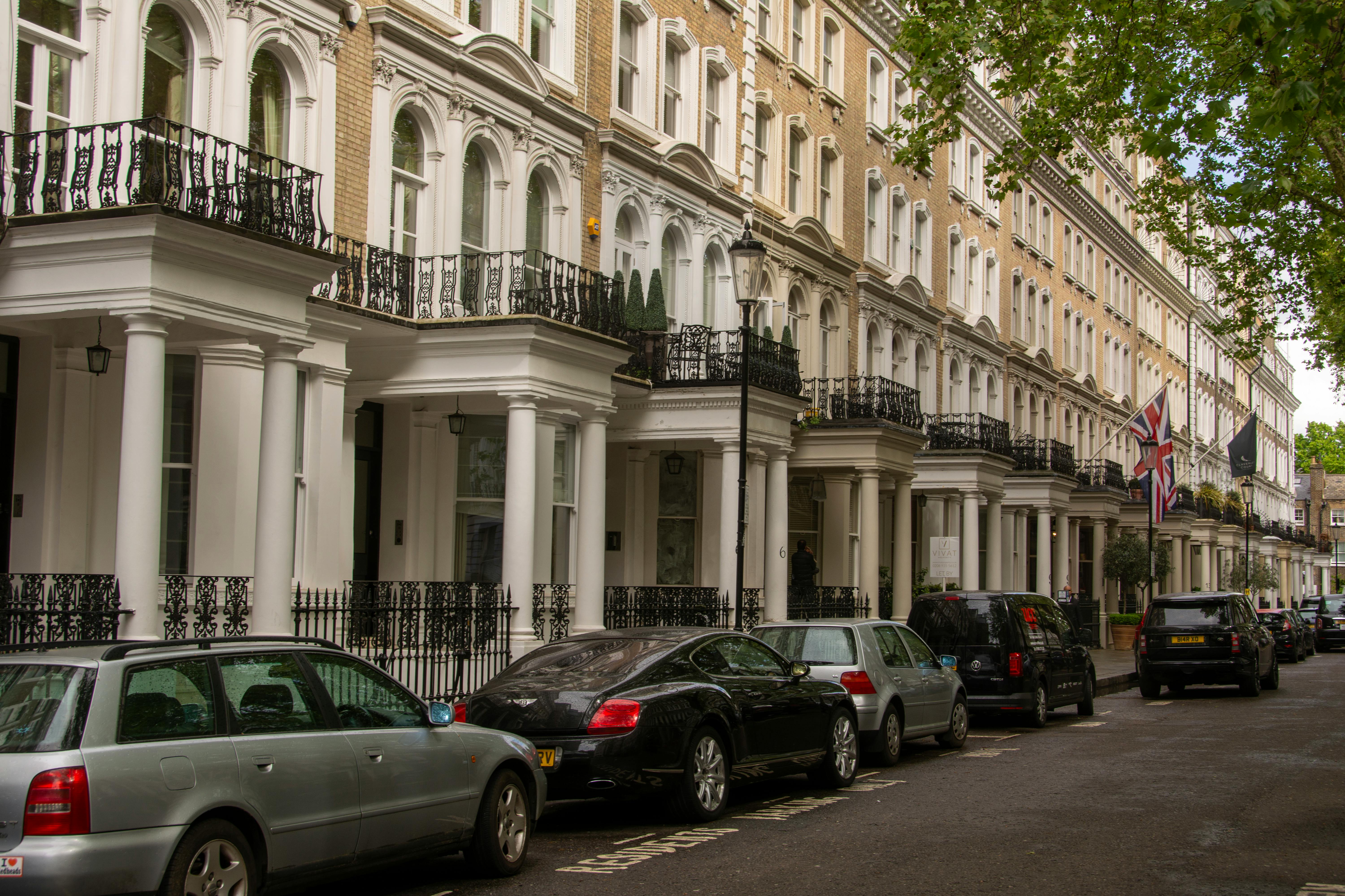 A row of multi-story terraced houses with classical architecture, featuring white columns, arched windows, and ornate iron railings. Union Jack flags hang from the buildings, and parked cars line the street. Green trees partially frame the scene, suggesting an upscale London neighborhood.