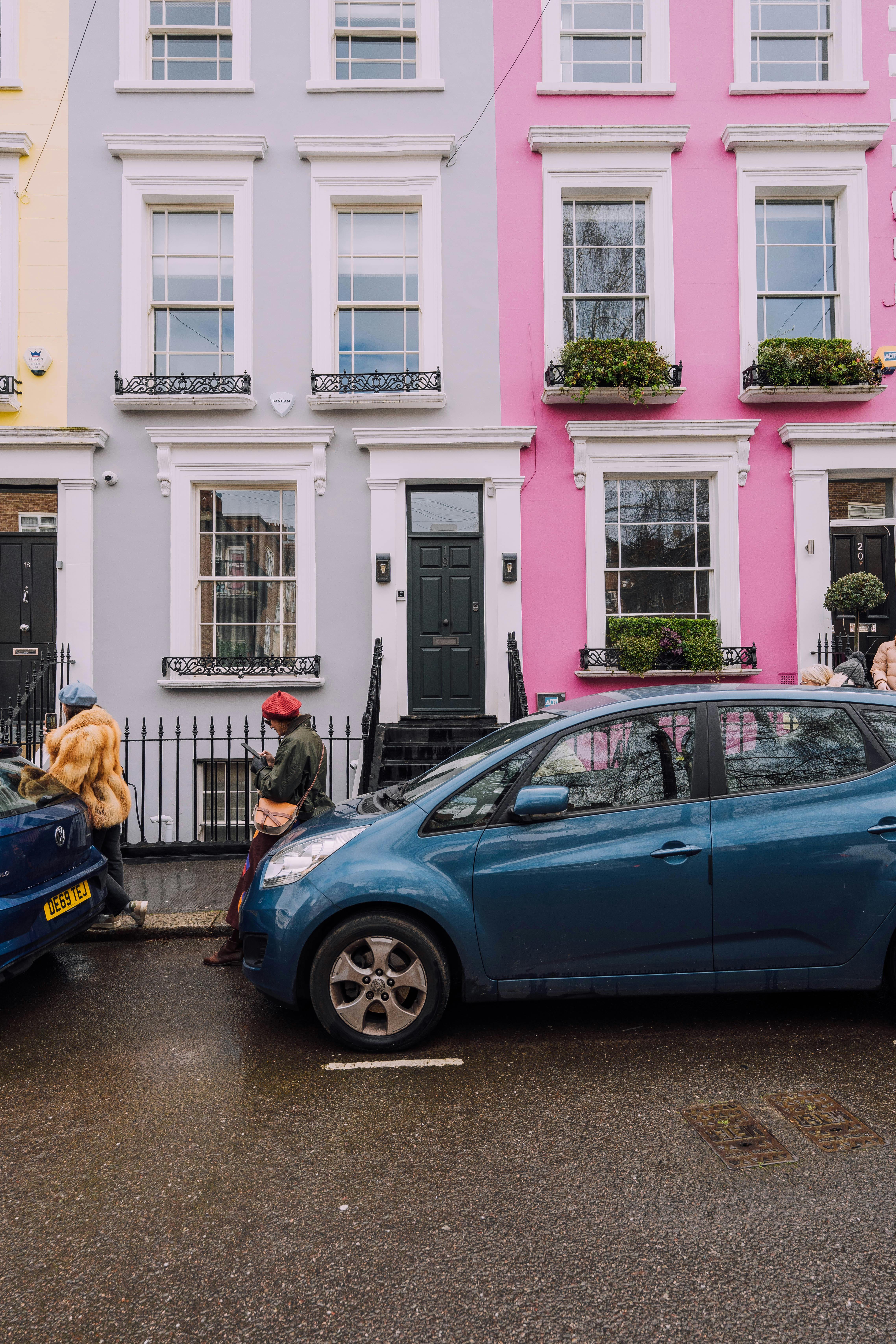 Pastel-colored townhouses in yellow, lavender, pink, and white line a quiet urban street. Each features large sash windows with white trim, black wrought iron railings, and dark-colored doors. Two stylish pedestrians walk along the sidewalk—one in a fur coat and blue hat, the other in a red beret and green jacket, looking at a phone. Two cars are parked nearby, one with a visible license plate reading “DE59 TEJ.”