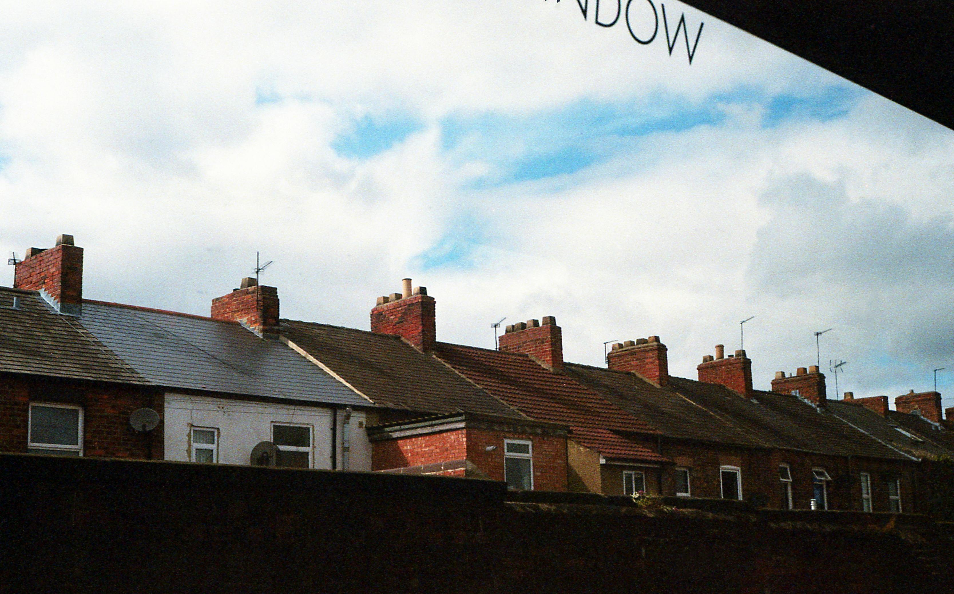 A row of closely packed terraced houses with red brick chimneys and sloped tiled roofs stretches across the frame. Some rooftops feature satellite dishes and TV antennas. The sky is partly cloudy with patches of blue, and the word “WINDOW” is faintly visible at the top, likely reflected or printed on the glass through which the photo was taken.