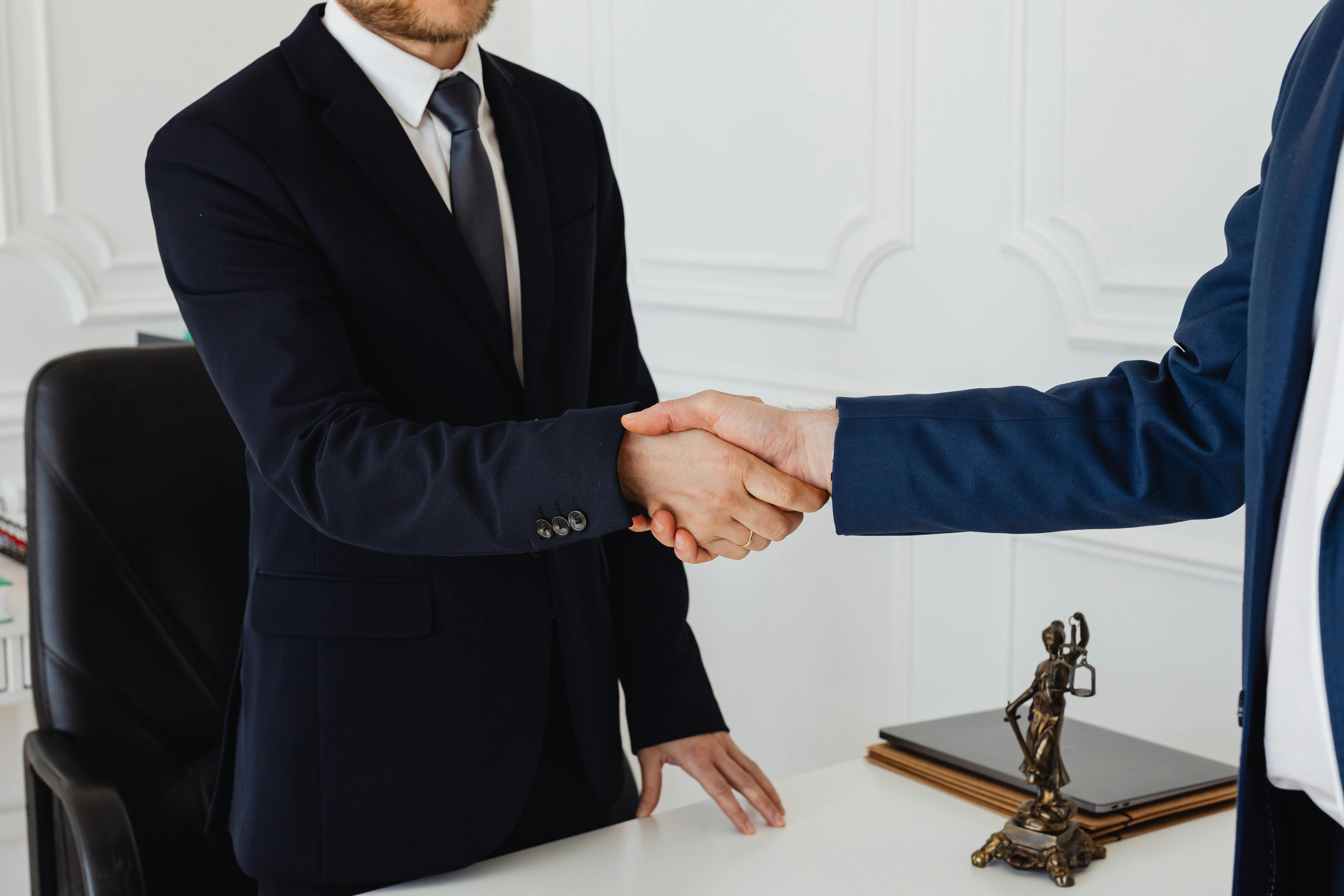 Two individuals in formal business attire shake hands in an office setting. One is seated behind a desk, while the other stands in front. On the desk are a laptop, a stack of folders, and a small statue of Lady Justice, reinforcing the legal or professional environment. The handshake suggests a formal agreement or conclusion of a business matter.