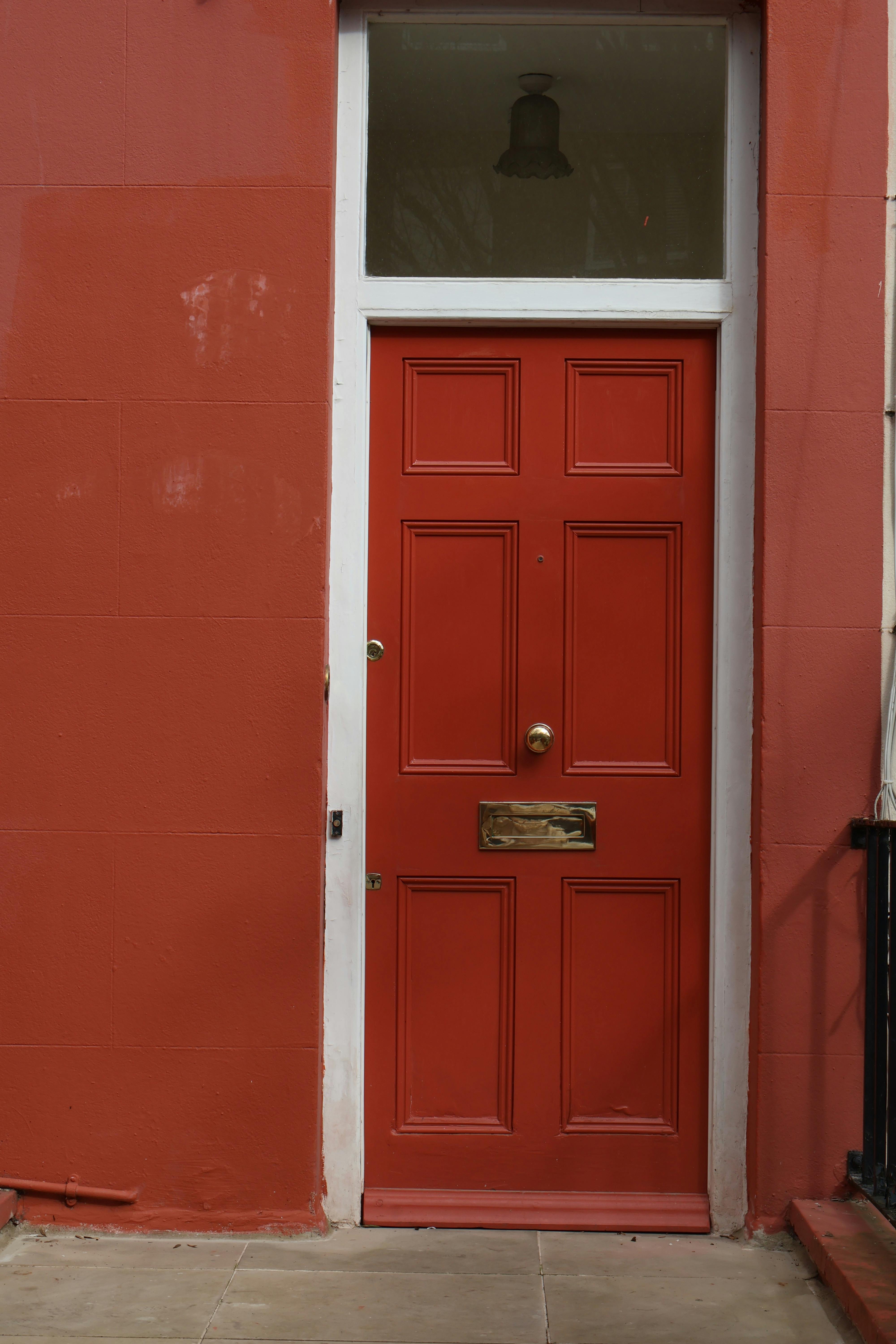A traditional red front door with six recessed panels is set into a red-painted exterior wall, framed by white trim. The door features a brass mail slot and doorknob, with a rectangular transom window above revealing an interior ceiling light. Light-colored stone tiles pave the ground in front, emphasizing the bold color contrast and classic architectural details.