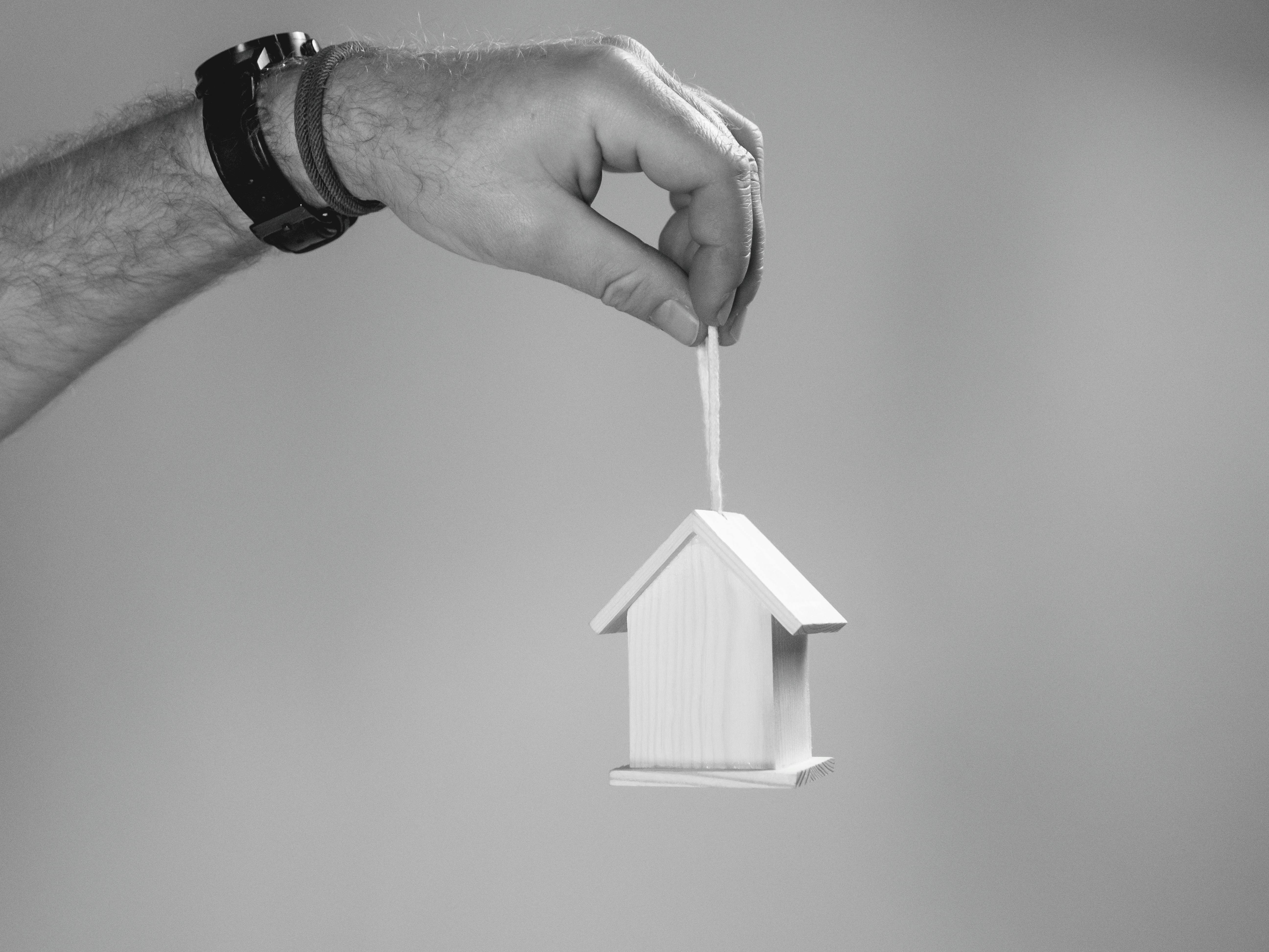 A hand wearing a wristwatch and bracelet holds a small wooden house model by a string. The house has a simple design with a triangular roof and rectangular base. The plain, softly blurred background draws focus to the symbolic gesture of homeownership or housing security.