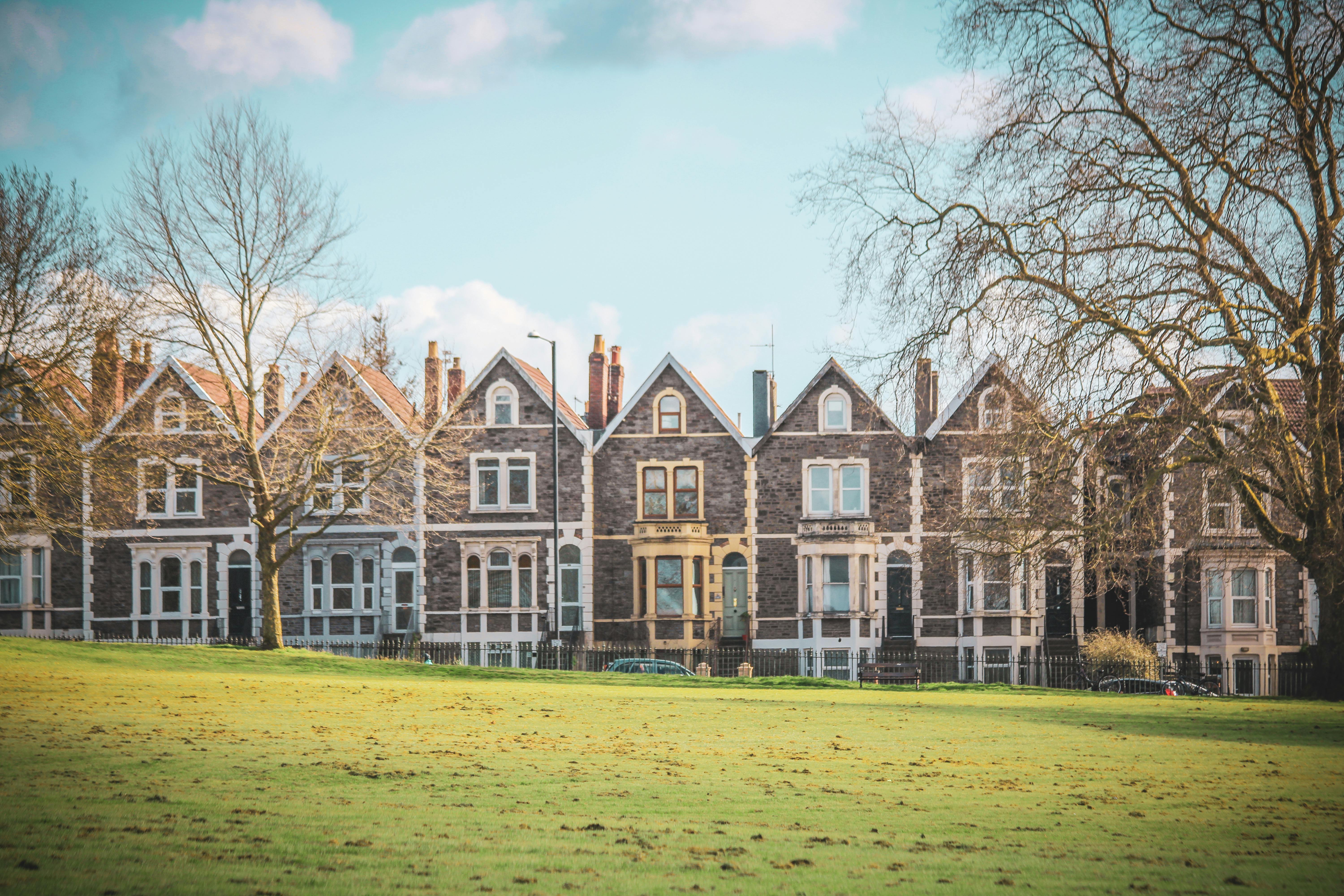 A row of Victorian terraced houses with dark stone facades, white trim, and ornate architectural details. Steeply pitched roofs, large sash windows, and decorative lintels add character. Leafless trees and a grassy field in the foreground suggest a winter or early spring setting under a partly cloudy sky.