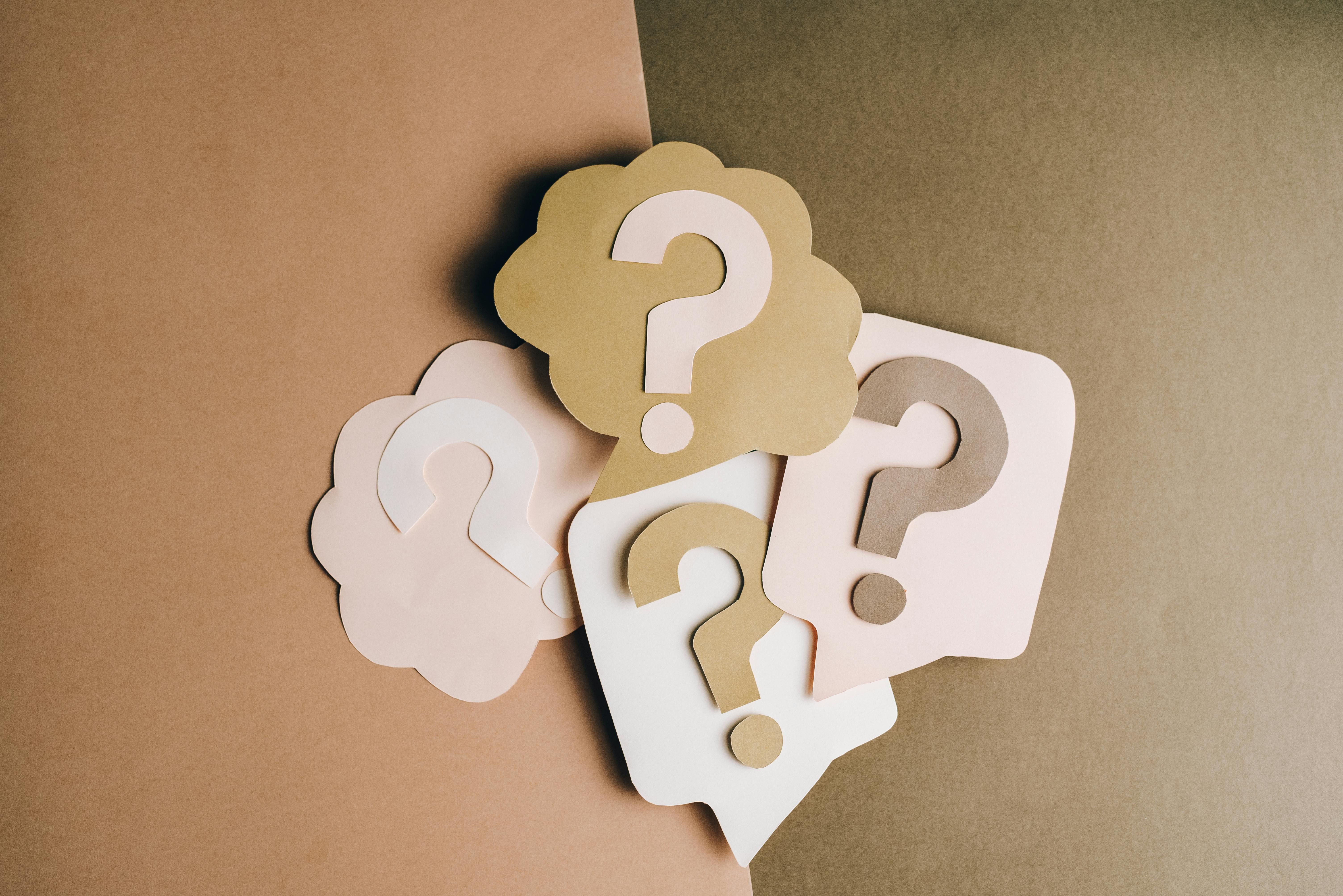 An arrangement of pastel-colored paper cutouts shaped like question marks placed on a two-tone brown background. The cutouts come in soft beige, cream, tan, and pink shades, with each having a unique outline style such as speech bubbles and cloud shapes.