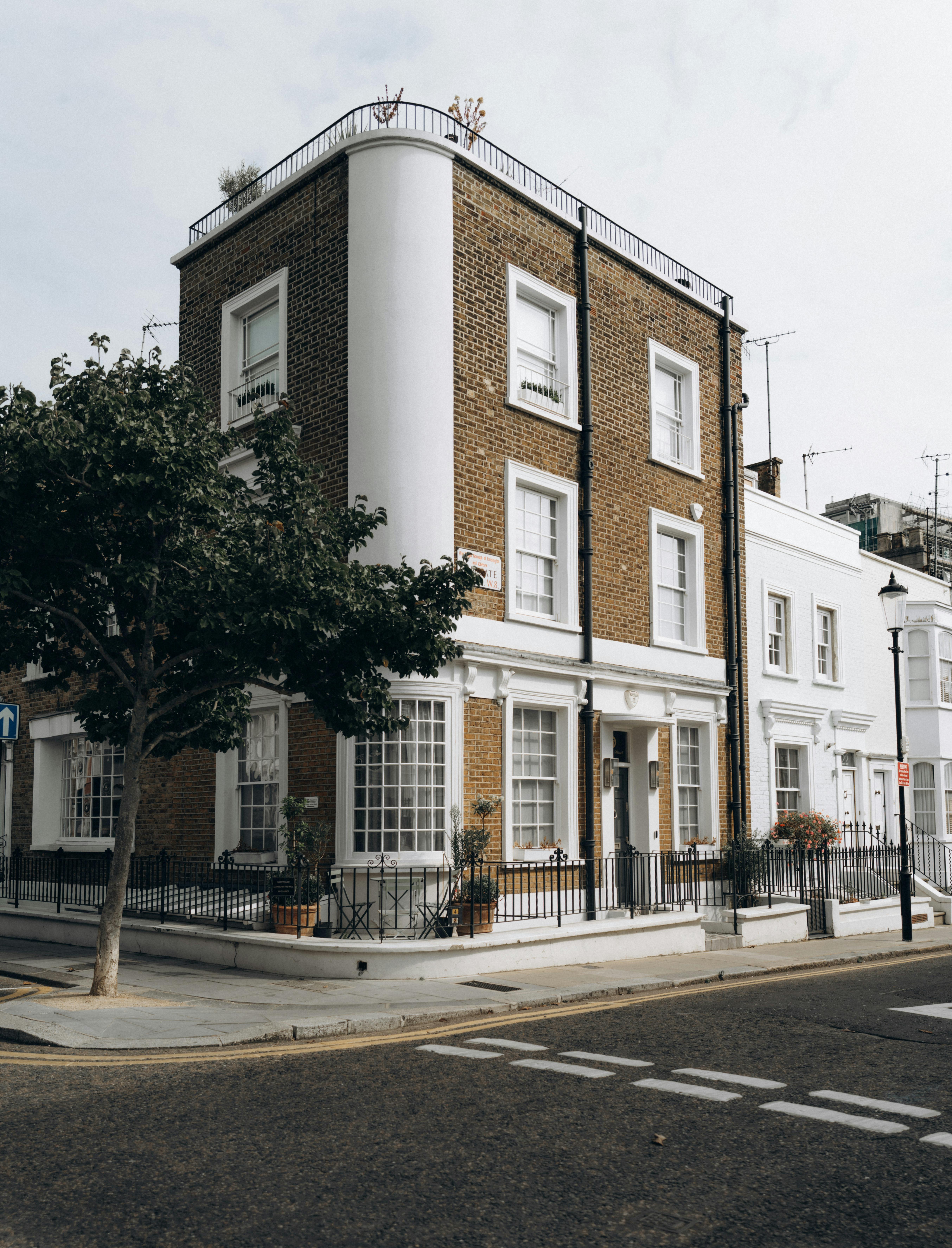 A three-story residential building with brown brickwork and white window accents stands at a quiet urban street corner. Black iron railings enclose the ground-level area, and potted plants decorate the windows and entrance. A tree grows on the sidewalk nearby, and road markings indicate a pedestrian crossing. The scene evokes classic European architecture and a peaceful city atmosphere.