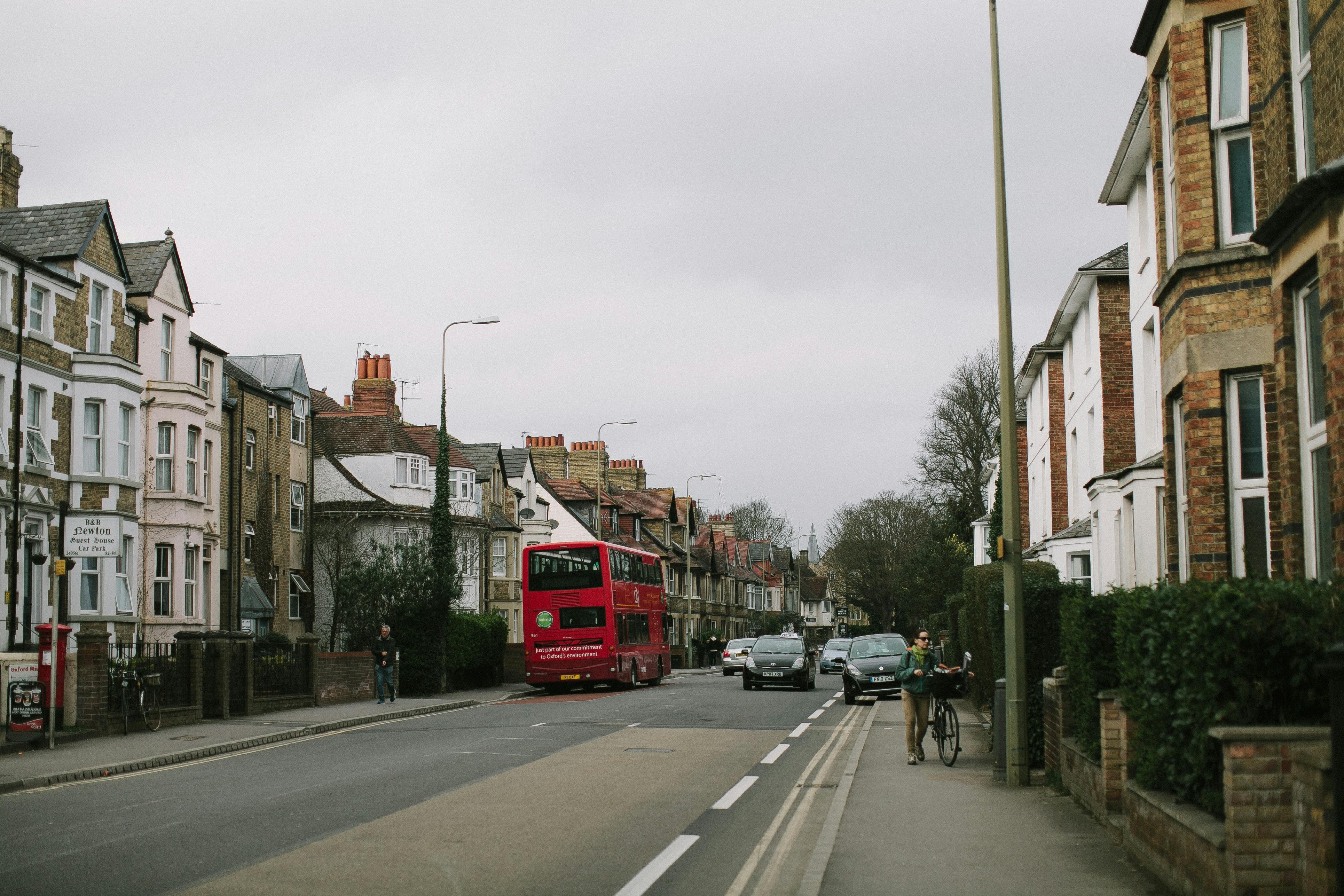 A residential street scene shows traditional brick and stone houses with bay windows and chimneys. A red double‑decker bus travels down the road alongside several cars. On the left, a sign for “B&B Newton Guest House En‑Suite Rooms” marks a bed and breakfast. One person walks on the sidewalk near the guest house, while another walks a bicycle on the opposite side. The road includes a designated bike lane, bordered by sidewalks and hedges. The sky is overcast, giving the scene a subdued atmosphere.
