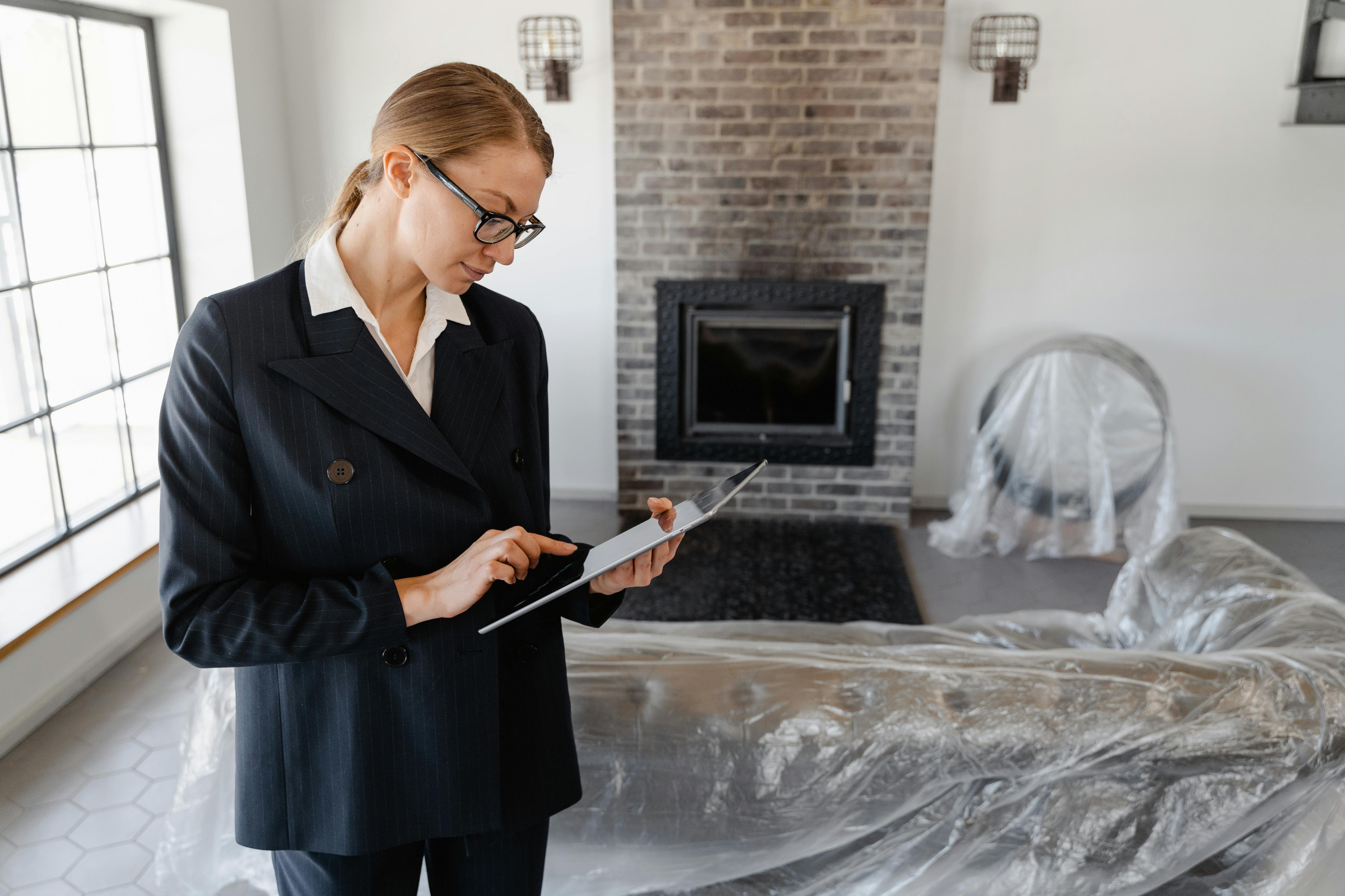 A person in a dark pinstripe suit with a white shirt holds and interacts with a tablet in a modern, minimally decorated room. The space features a brick fireplace, large black‑framed windows, hexagonal floor tiles, and wall‑mounted light fixtures. Furniture including a chair and couch is covered with protective plastic sheets, suggesting renovation or staging. The scene conveys a professional conducting an inspection or assessment in a residential setting.