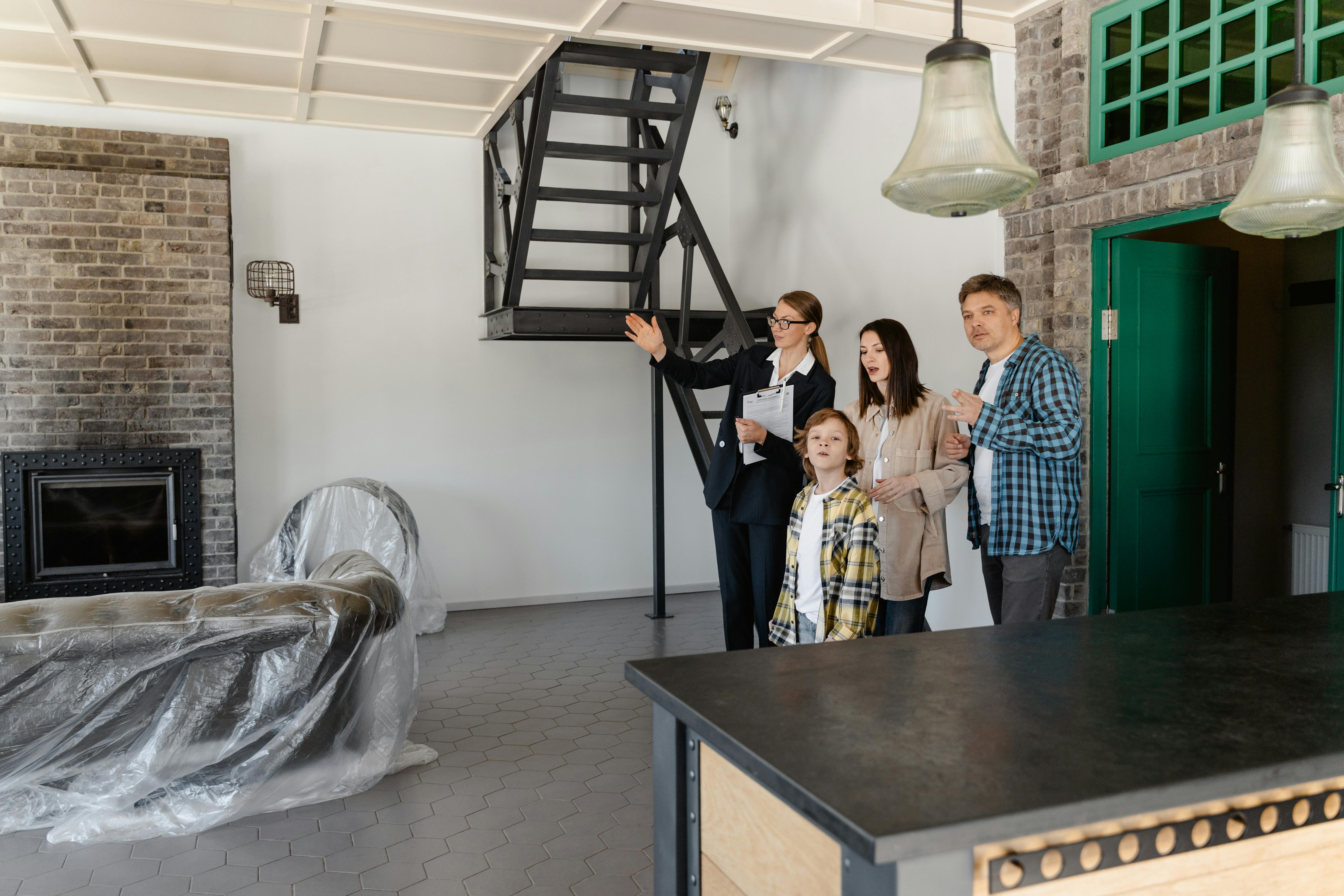 Inside a modern, industrial-style home, a professionally dressed woman gestures while holding documents, likely guiding a family of four through a property tour. The room features a brick wall with a fireplace, a black metal staircase, and furniture covered in plastic. Hexagonal floor tiles and large hanging lights add to the contemporary aesthetic.
