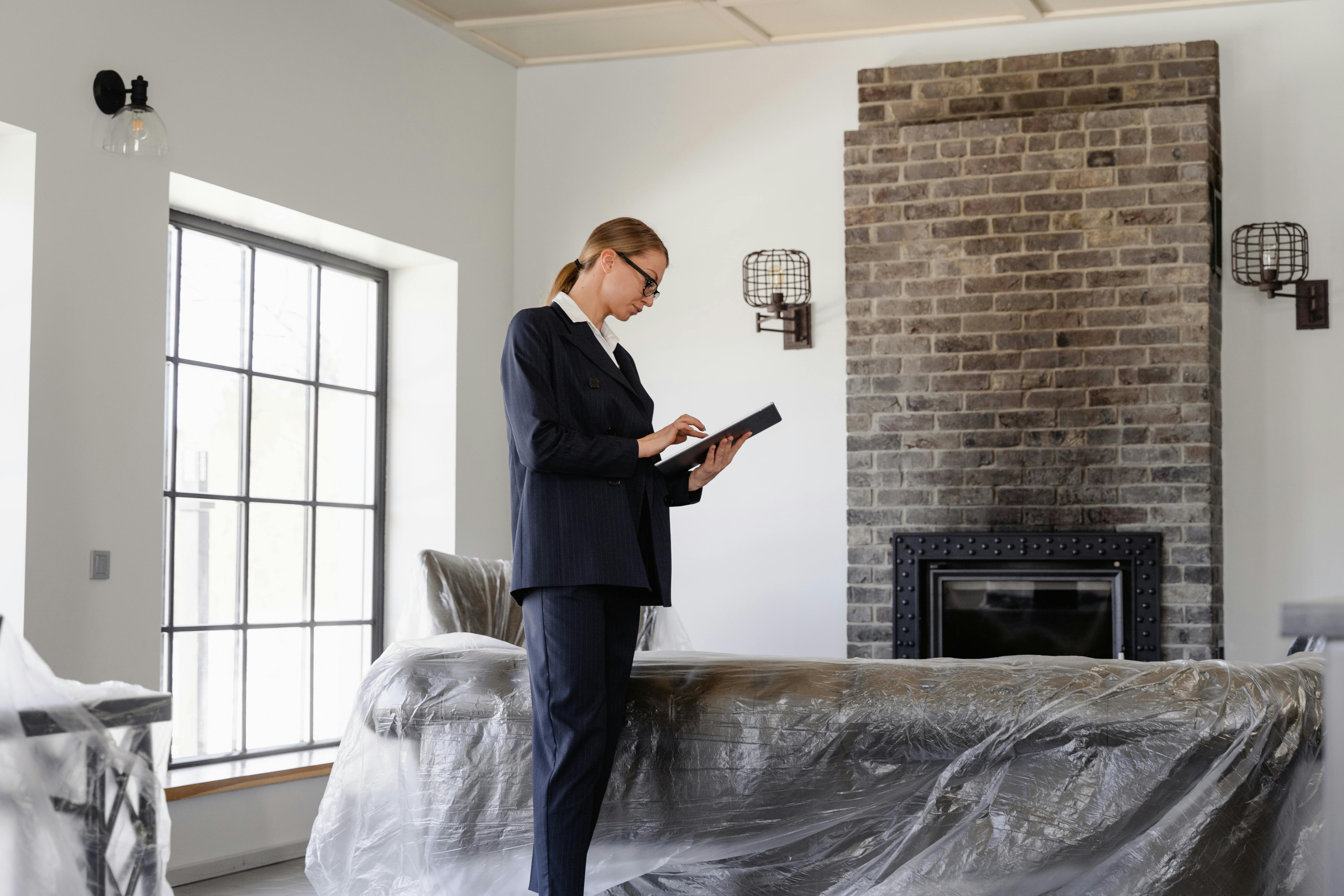 A person in a dark business suit stands in a modern living room, interacting with a tablet. The space features a large brick fireplace flanked by wall-mounted lights, a wide black-framed window, and furniture covered in protective plastic sheets, suggesting renovation or staging. Natural light fills the clean, contemporary room.