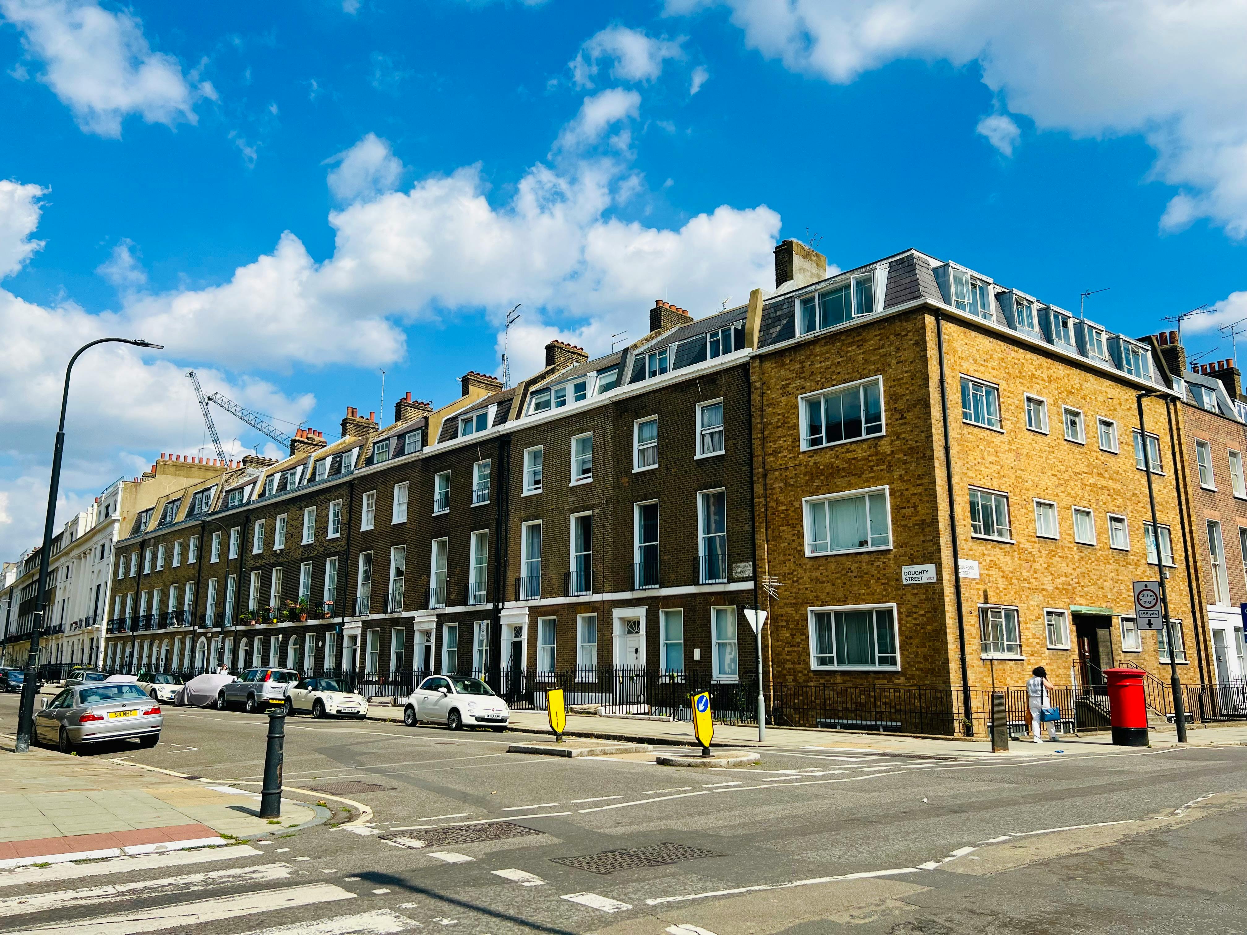 A street corner in a British urban neighborhood features attached brick buildings with large windows and dormers. The corner building, made of lighter bricks, displays a “Doughty Street” sign. Parked cars, a pedestrian, and a red post box add life to the scene. A street sign restricts heavy vehicles, and a construction crane in the background hints at ongoing development under a bright, partly cloudy sky.