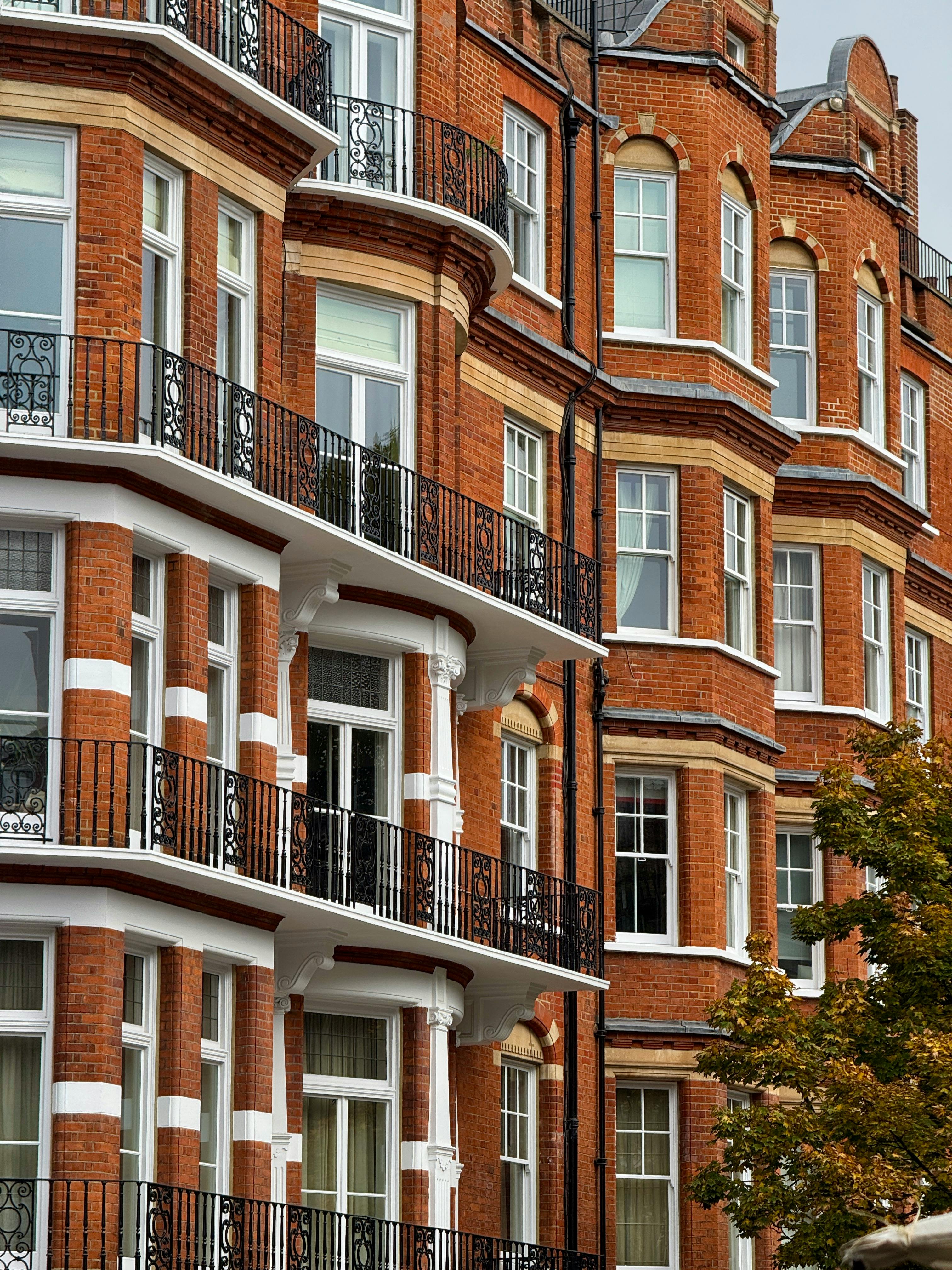 A red-brick urban residential building features white-framed windows, ornate black wrought iron balconies, and curved bay windows. Decorative cornices and symmetrical window placement suggest Victorian or Edwardian architectural influence. A tree with green and yellowing leaves in the foreground hints at early autumn, adding warmth and texture to the historic facade.