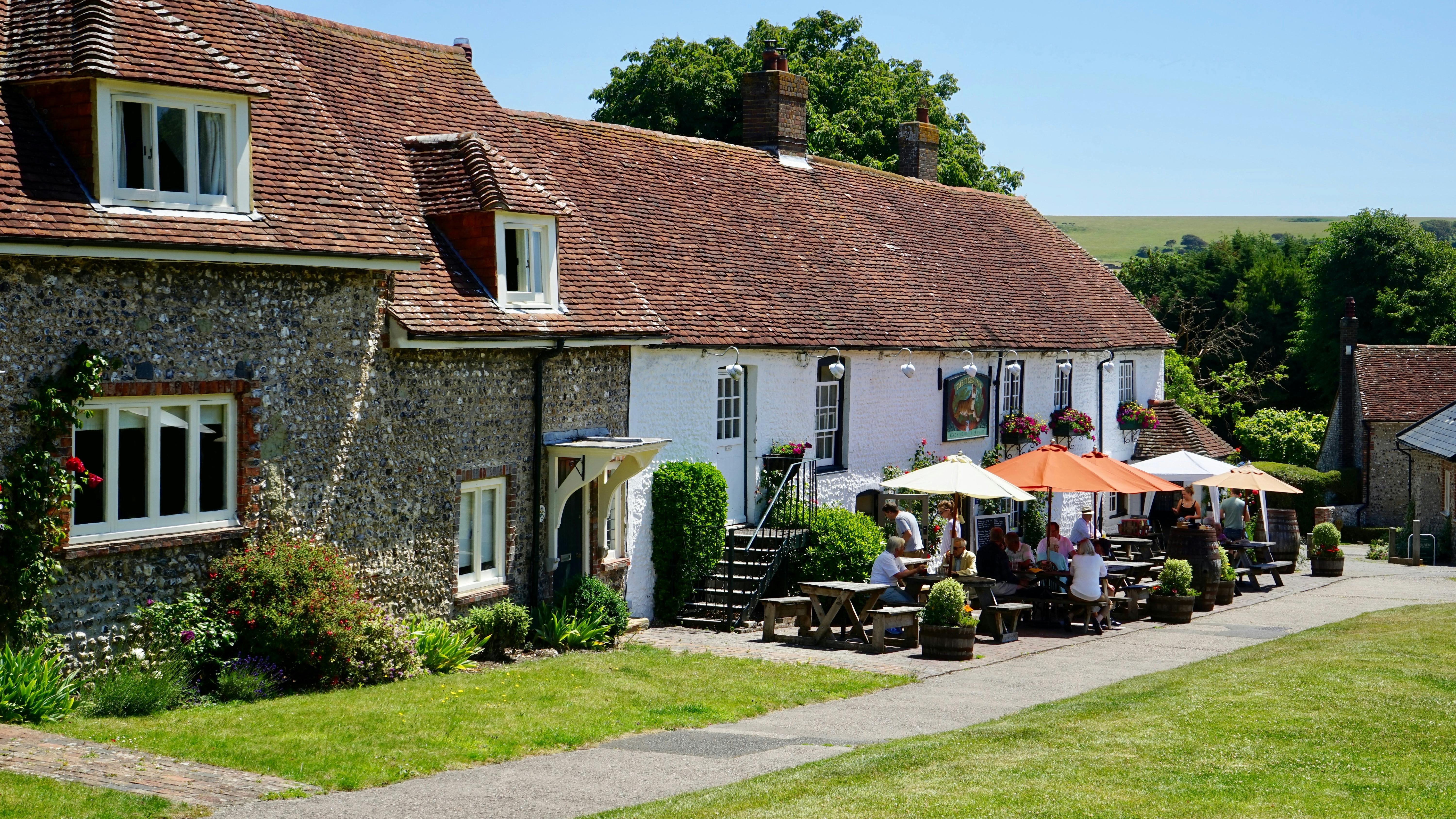 A traditional English-style stone building with a red-tiled roof serves as a countryside pub or restaurant. Flower boxes and hanging baskets filled with vibrant blooms decorate the exterior. Groups of people sit at wooden picnic tables under large umbrellas, enjoying food and drinks in a lush garden setting. A signboard with an illustration hangs on the white-painted section of the building, and trees and greenery frame the peaceful rural scene.