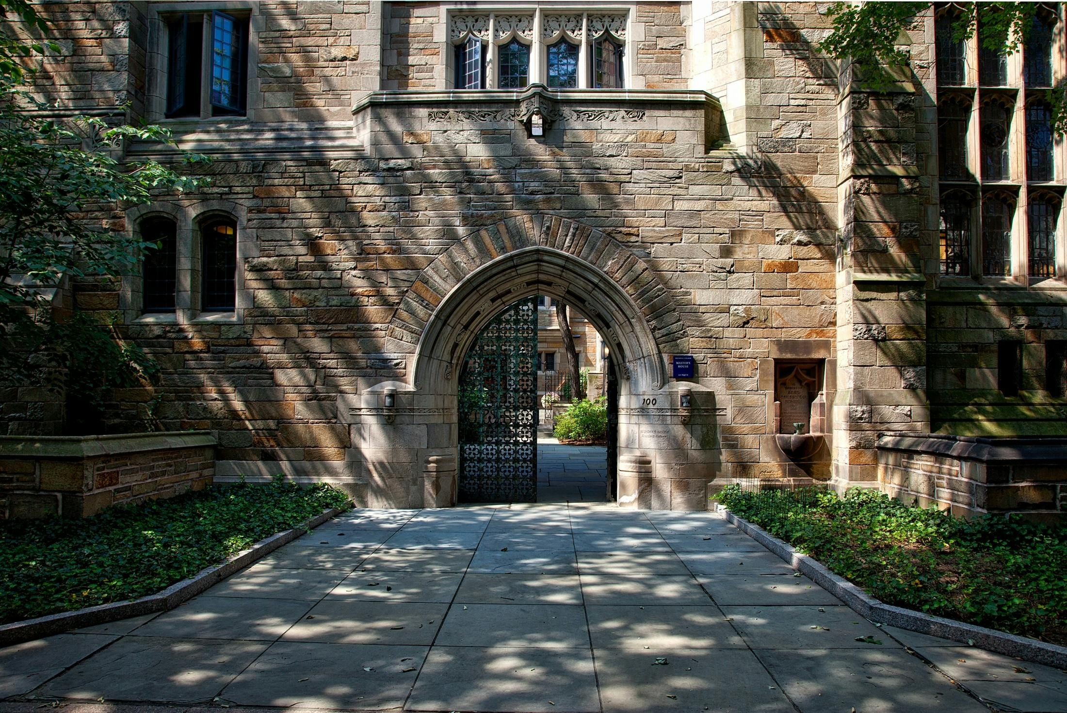 A stone building with Gothic architectural design featuring an arched doorway, ornate carvings, and decorative windows. The arched entrance is partially open, revealing a pathway leading to a garden courtyard beyond. The exterior walls are built with textured tan and gray stone, and sunlight filters through nearby trees, casting dappled shadows across the walkway and facade.