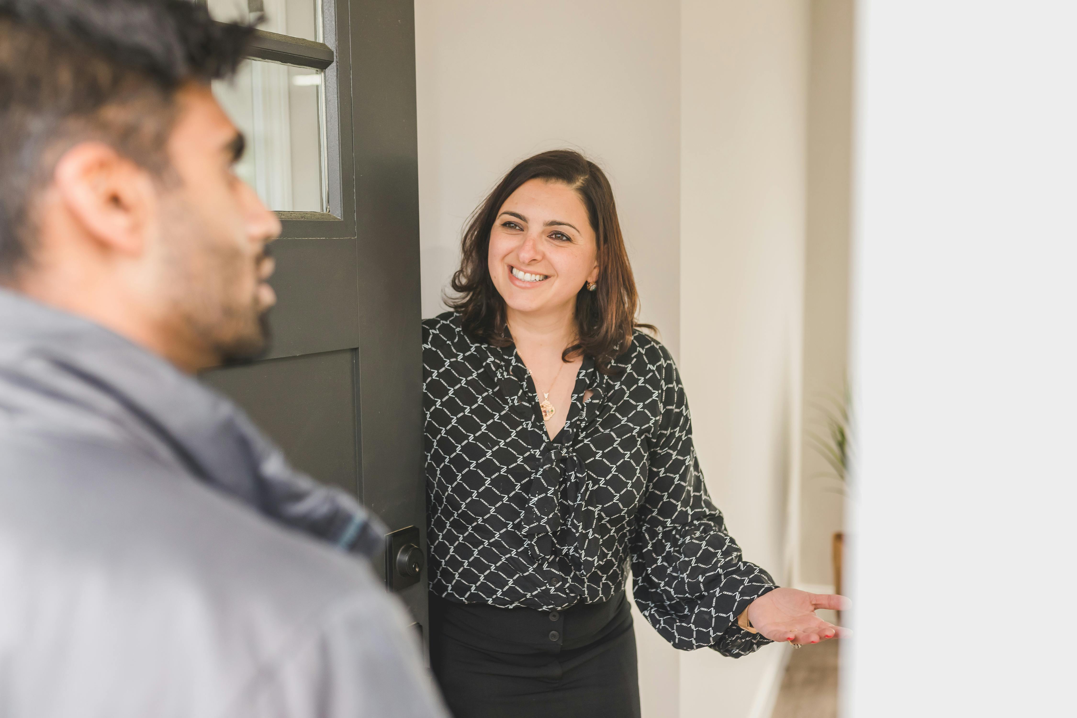 A woman in a black blouse with a white geometric pattern and black skirt stands at the entrance of a home, smiling and gesturing toward a man outside the door. The man, partially visible in a gray jacket, appears engaged in conversation. The open door reveals a light-colored interior wall and a potted plant, suggesting a warm, welcoming interaction.