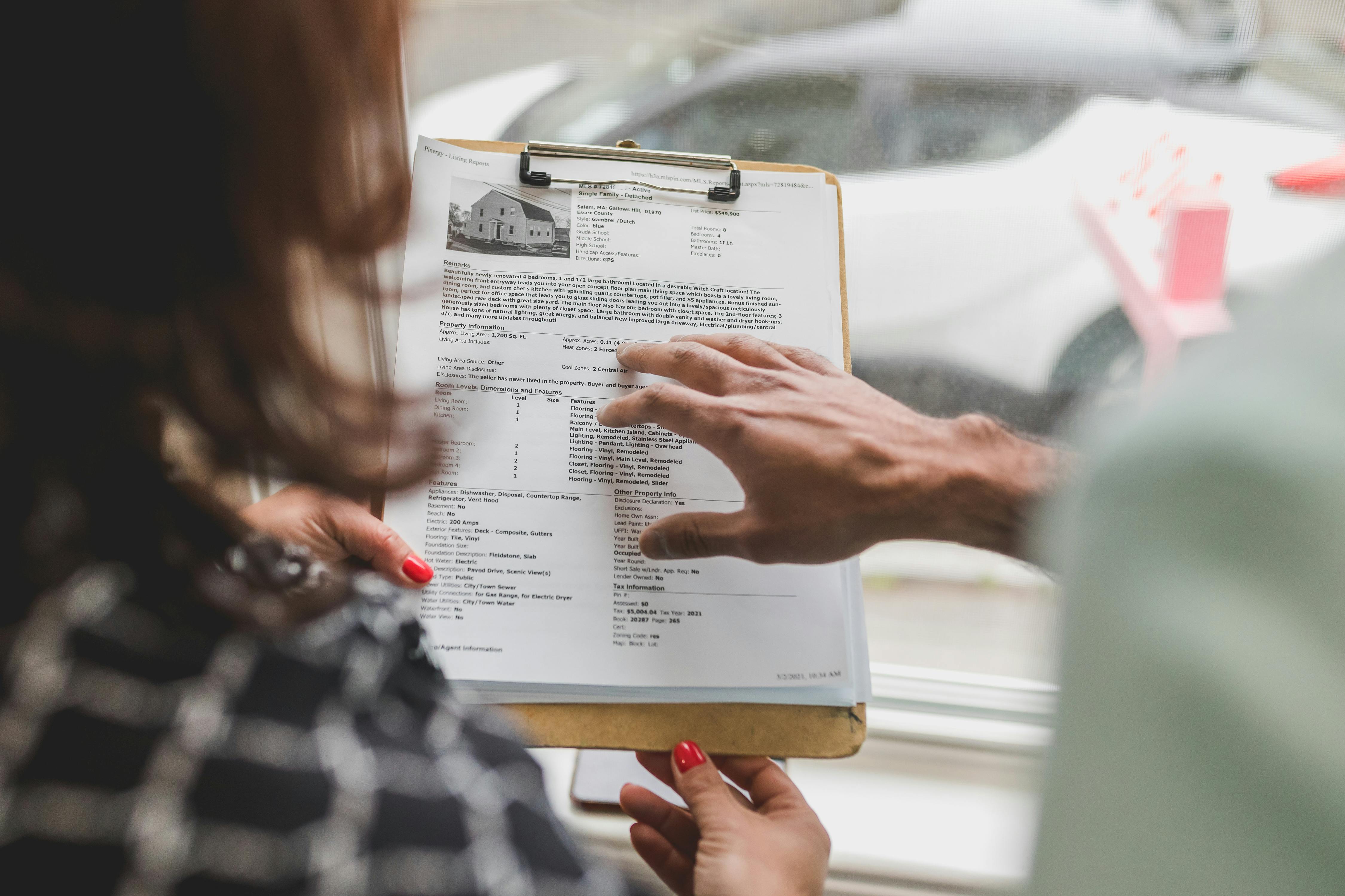 Two individuals review a property listing document on a clipboard. One person holds the clipboard while the other points to the page, which includes a photo of a house, the address “123 Main Street, Anytown, MA,” and detailed real estate information such as price, features, room dimensions, and amenities. The scene suggests a home tour or real estate consultation.