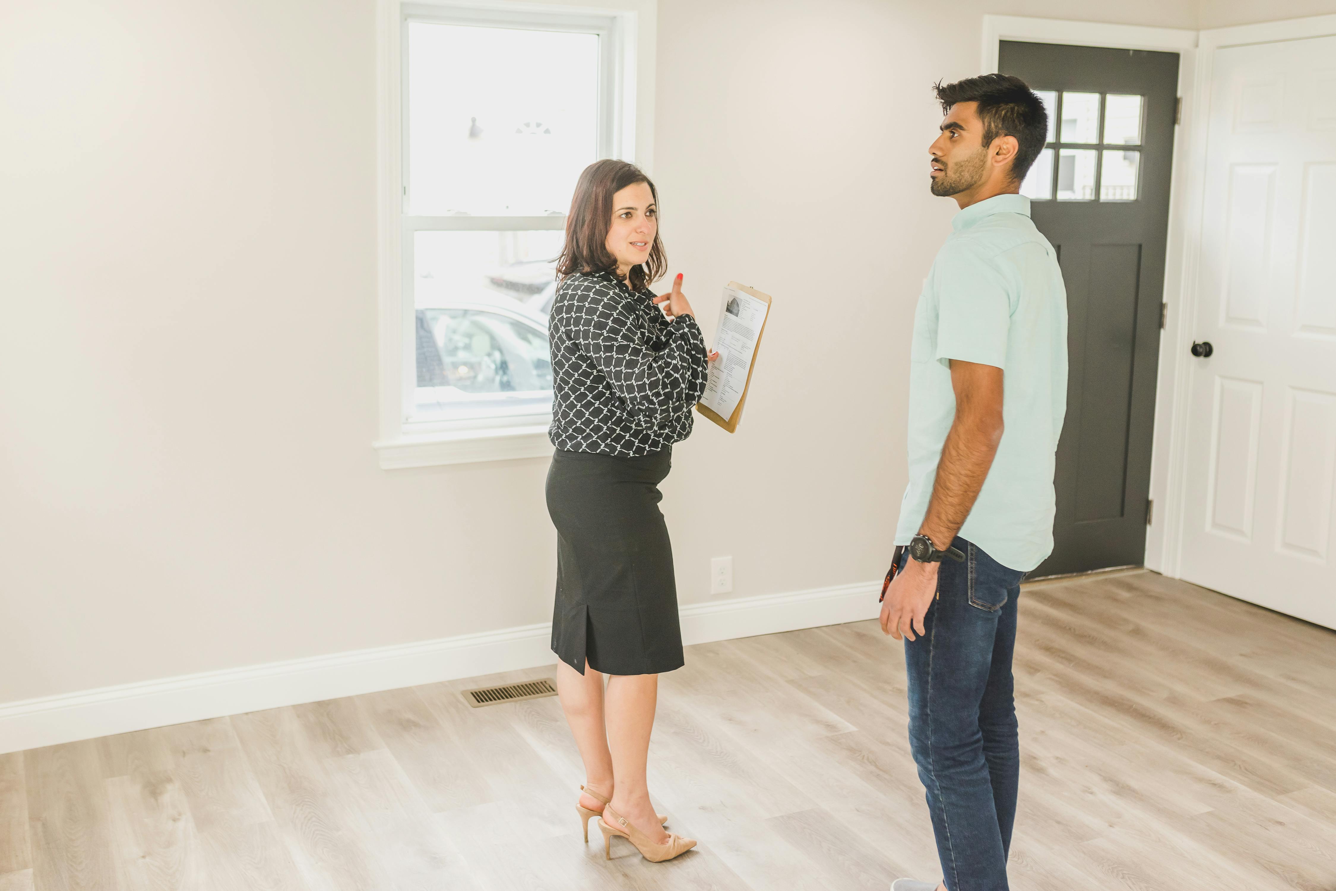 Two individuals stand in a light-colored room with wood flooring, engaged in discussion. One holds a clipboard with printed documents, possibly related to property inspection or real estate. A window reveals a parked car outside, and a black door with glass panels adds contrast to the clean, modern interior.