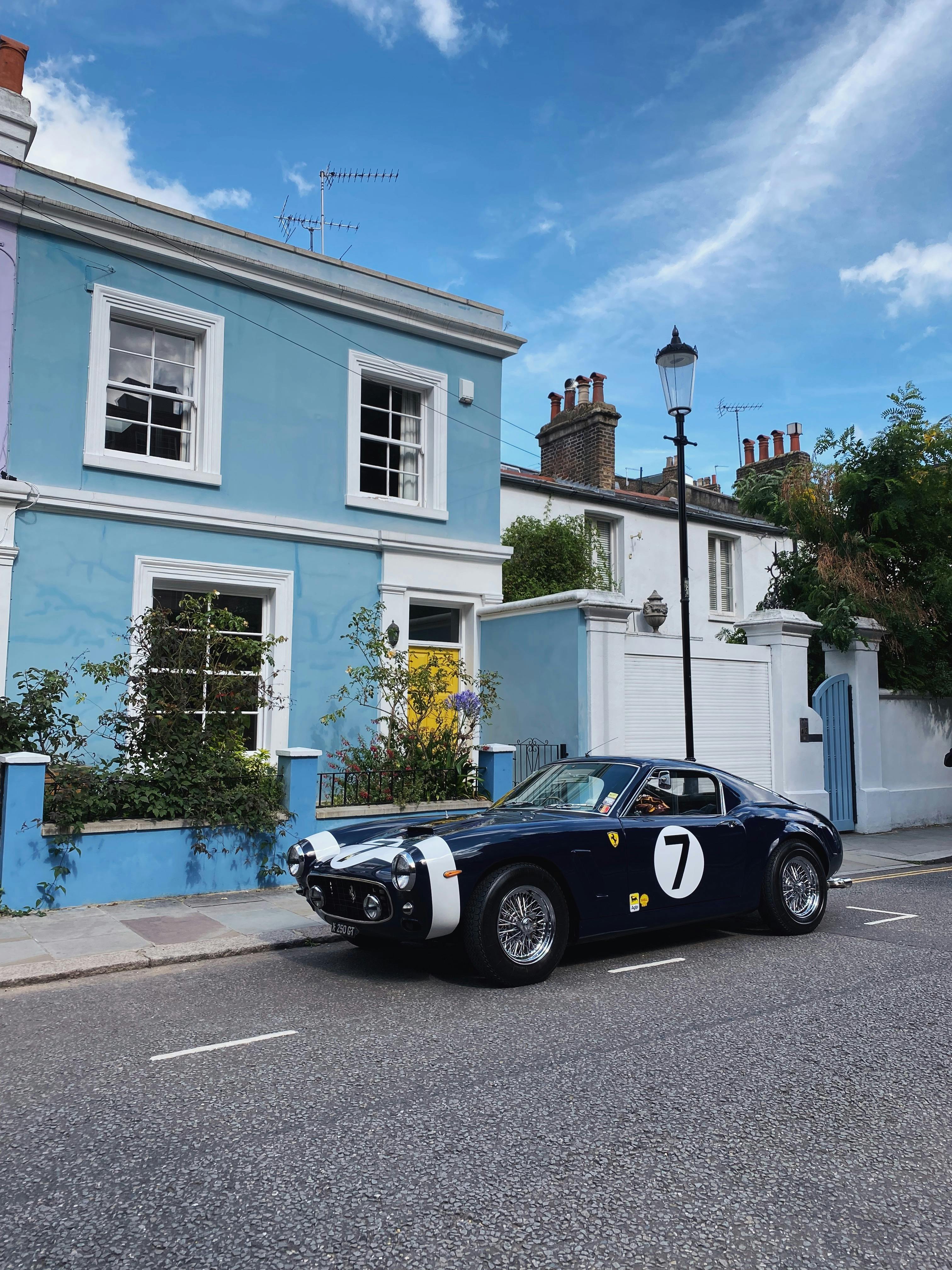 A dark blue vintage Ferrari with white racing stripes and the number “7” on the door is parked on a quiet residential street. The car sits in front of a pastel blue house with white trim and a bright yellow front door, surrounded by greenery. A traditional black streetlamp and similar houses line the background under a clear sky with wispy clouds.