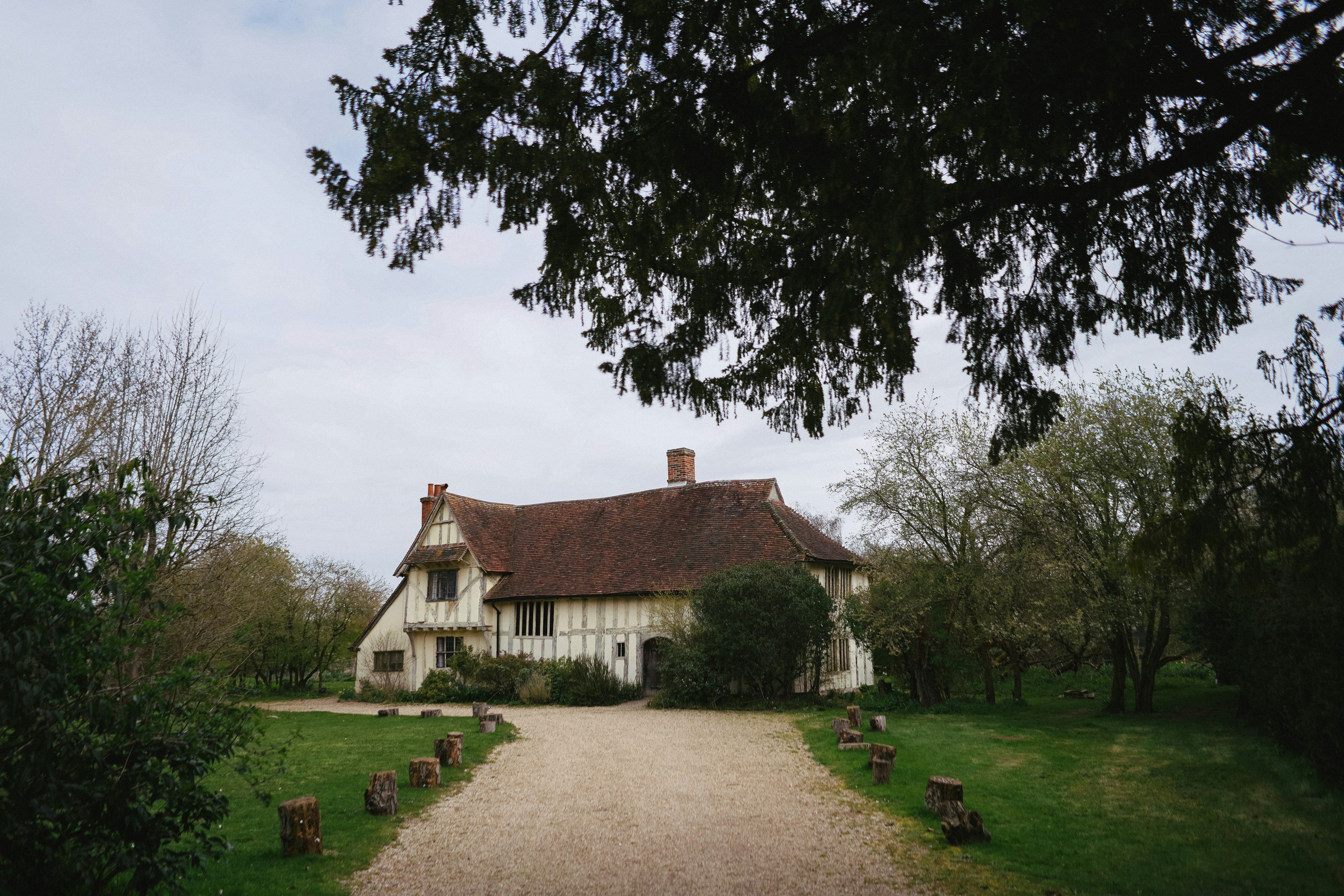 A picturesque house with timber-framed walls and a steep red-tiled roof, set amid lush greenery. A gravel driveway leads to the entrance, flanked by evenly spaced tree stumps. The home features dormer windows, a central chimney, and a tranquil garden setting framed by overhanging tree branches.