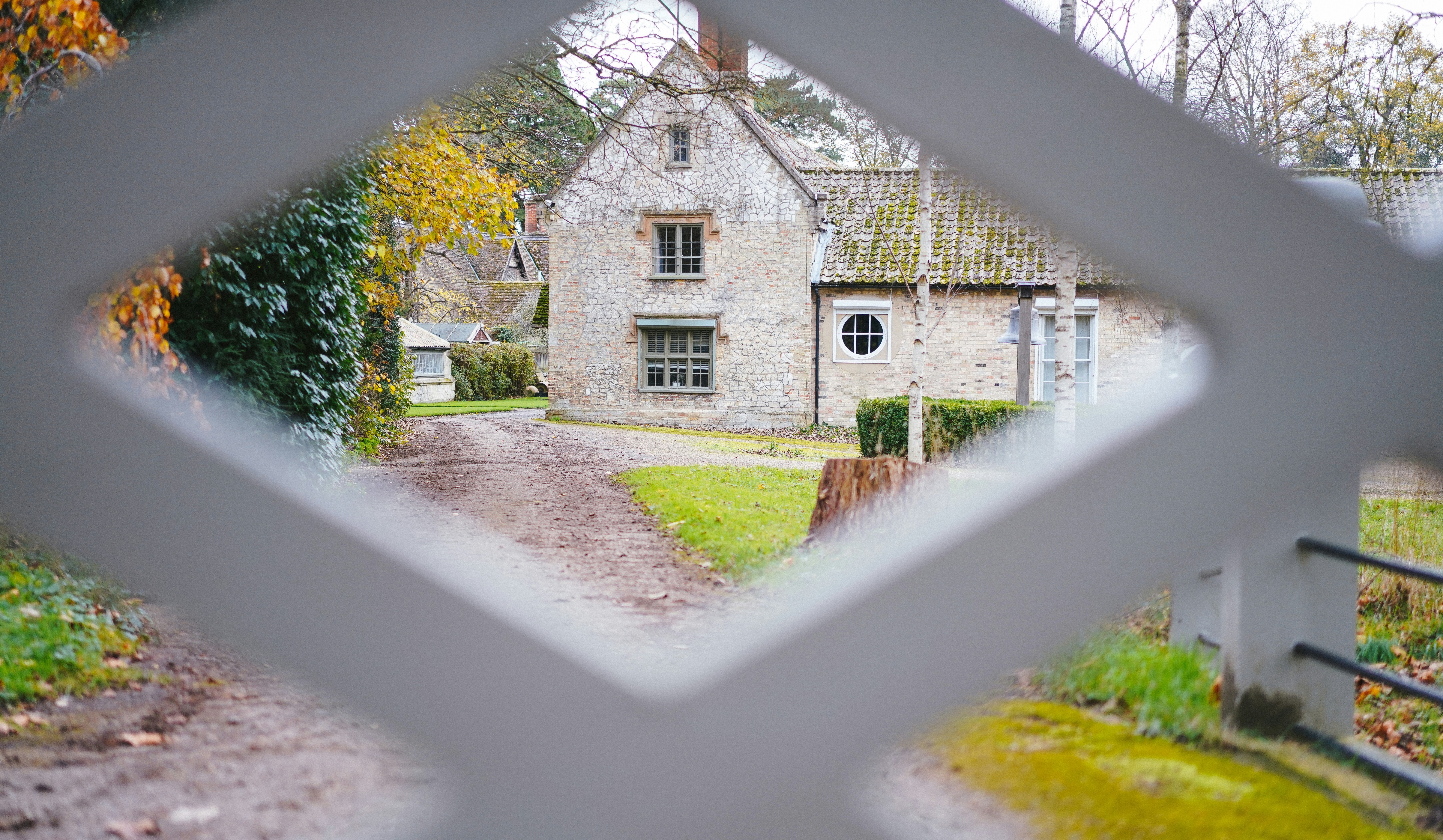 A charming stone house with a tiled roof and circular side window is viewed through a diamond-shaped opening in a white fence. A dirt path leads to the house, bordered by green grass and trimmed hedges. Surrounding trees with autumn leaves and bare branches suggest a fall setting, while the fence framing adds depth and visual interest.