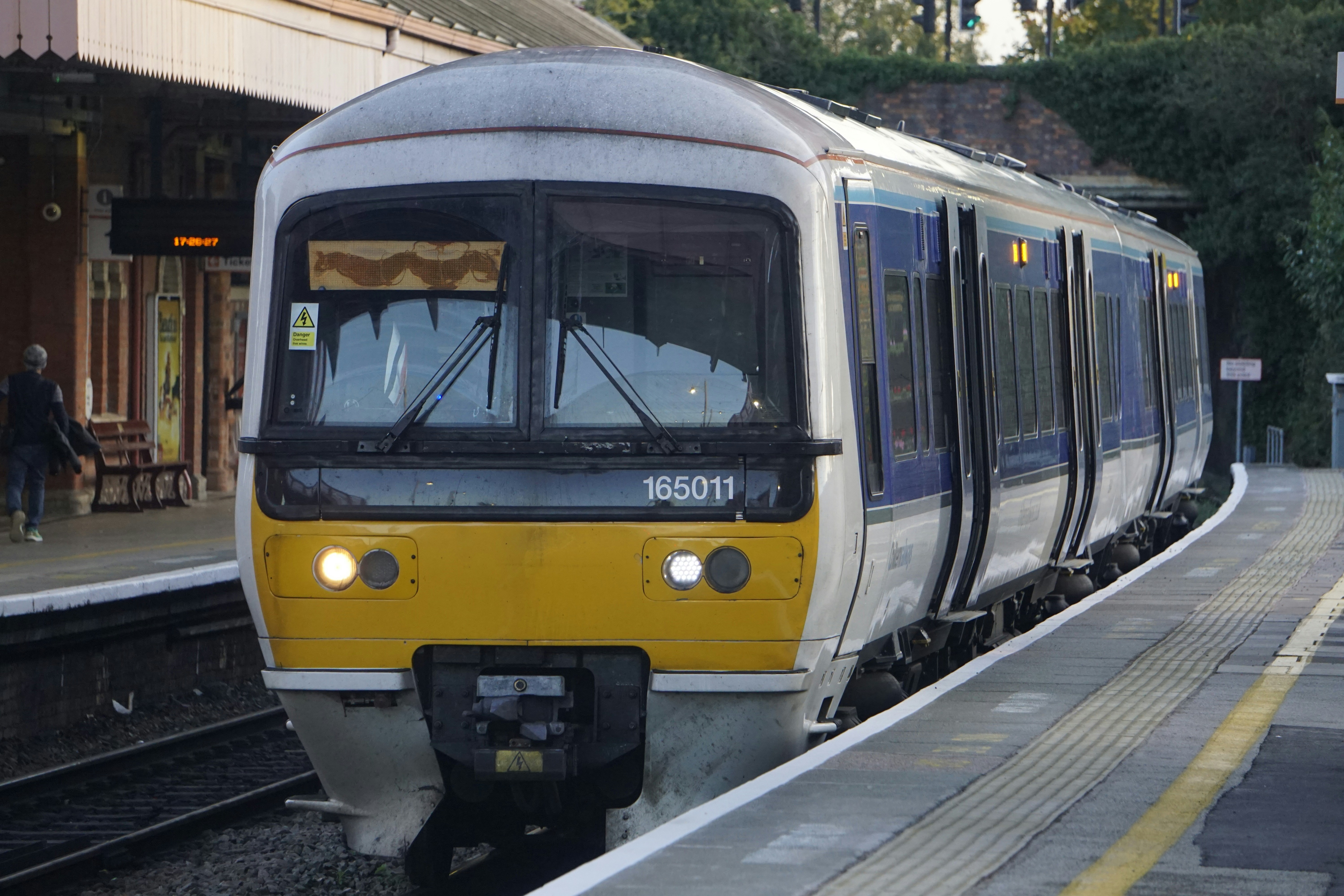 A Chiltern Railways passenger train with a yellow front marked “165011” is stationary at a platform. A person walks nearby, and a digital clock reads 17:08:07. The station features traditional brickwork and a canopy, capturing a moment of public transport in use.