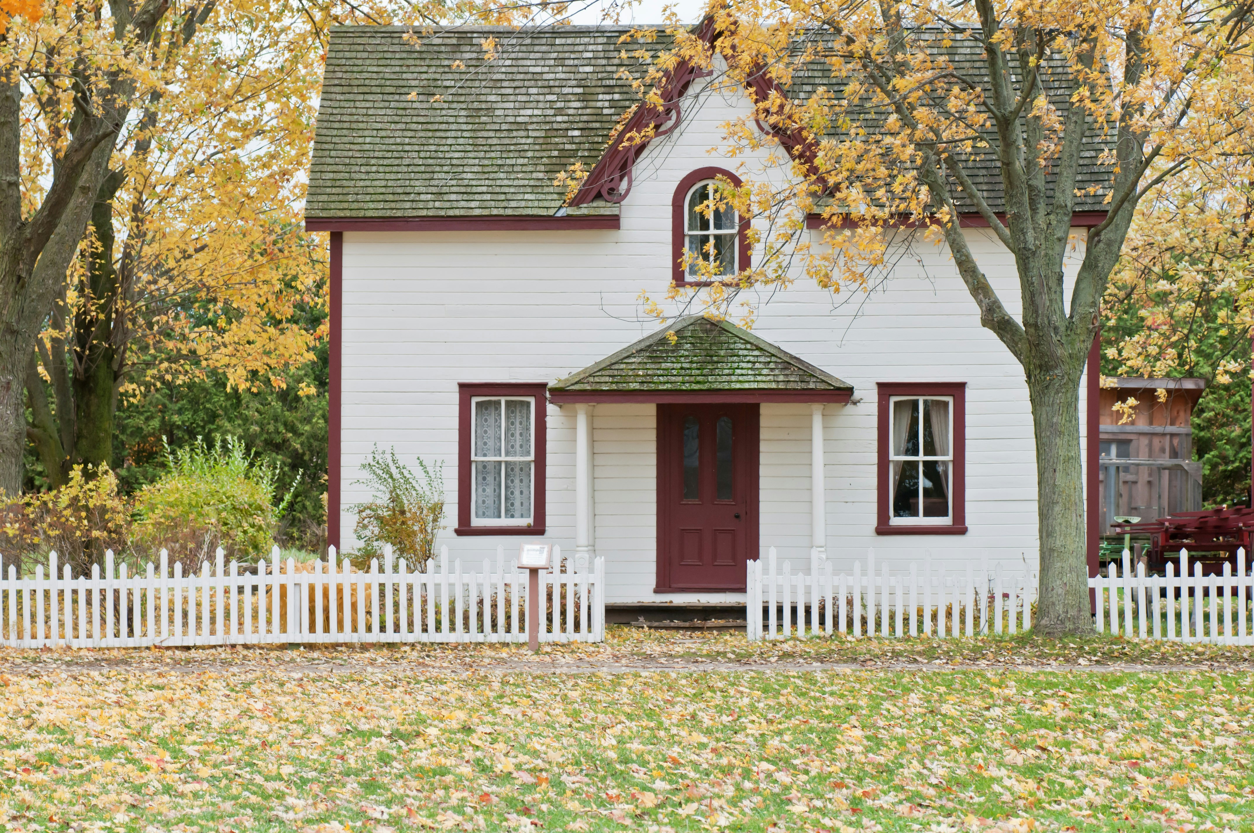 A quaint white two-story house with red trim and a steeply pitched roof stands behind a white picket fence. The symmetrical facade features a central front door, two windows, and a small gabled porch supported by white columns. Fallen leaves cover the yard, and mature trees with yellow foliage frame the scene, evoking a serene autumn atmosphere.