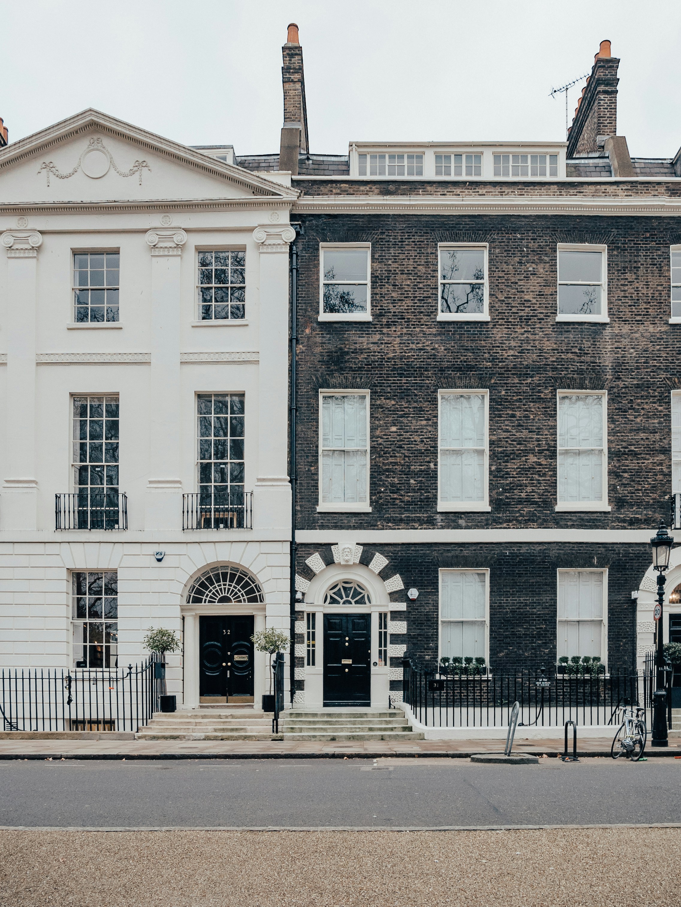 Two adjacent Georgian-style townhouses stand side by side in an urban setting. The left building is painted white with classical columns, moldings, and a pediment, while the right features a dark brick facade with white-trimmed arched windows and a decorative entrance. Both have symmetrical window arrangements and black front doors with brass fixtures. A bicycle, sidewalk, and traditional streetlamp complete the clean streetscape.