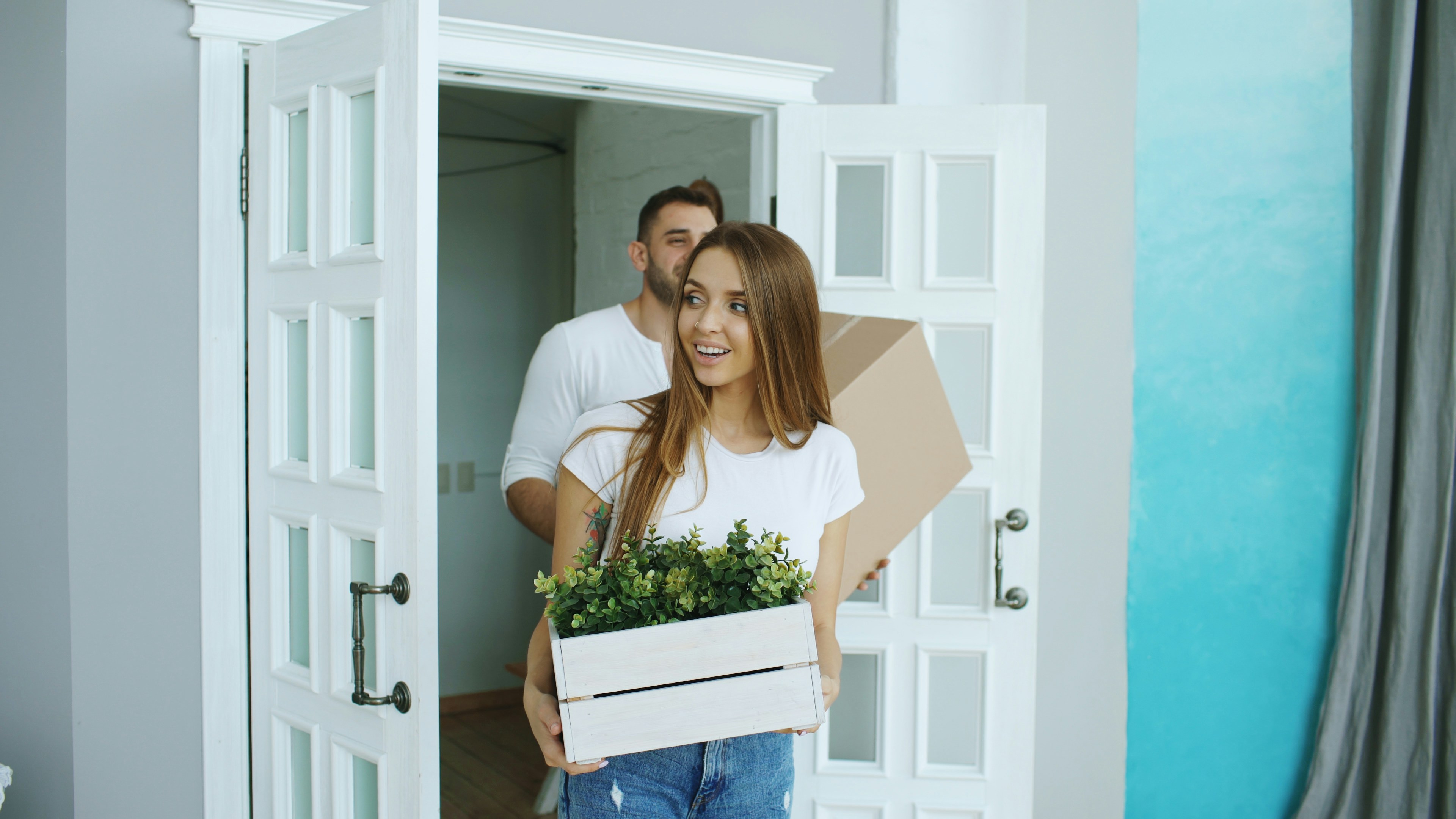 Two casually dressed individuals enter a room through a white door with glass panels. One carries a white wooden box filled with green plants, while the other holds a large cardboard box. The room features light-colored walls, a blue gradient accent wall, and a curtain to the side, suggesting a fresh move or home setup in a bright, welcoming space.